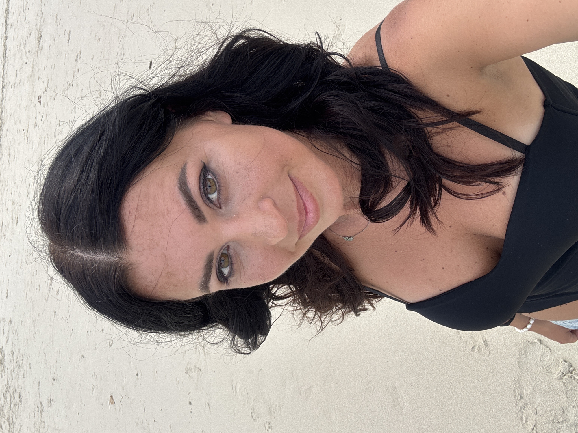 A woman is on a beach in Ban Haad Rin, Thailand. The woman has black hair and fair skin, wearing a black strappy top. The background is white sand with visible footprints.