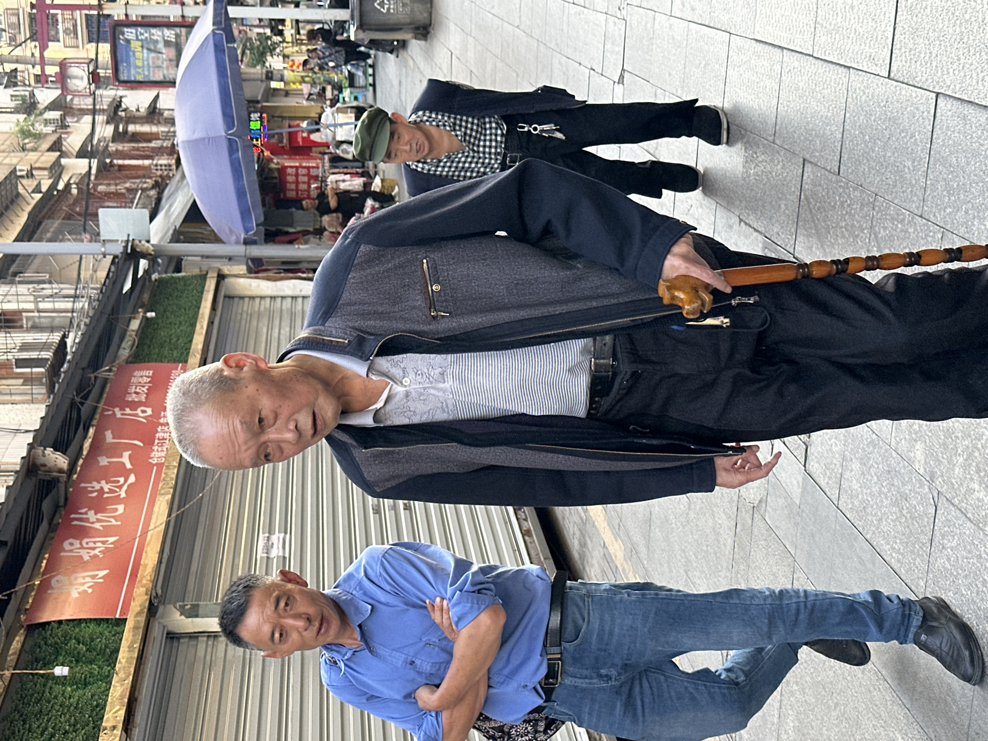 The image shows three middle-aged or older men standing on a sidewalk in an urban environment. One man is wearing a blue button-down shirt and jeans and has his arms crossed. Another man in the foreground is holding a walking stick and wearing a dark jacket over a light-colored shirt. A third man, further in the background, wears a green cap and a patterned shirt with a dark jacket. The background includes buildings with Chinese signage, a blue umbrella, and other pedestrians. The signage is written in Chinese and indicates the location is Jiangjin, China.