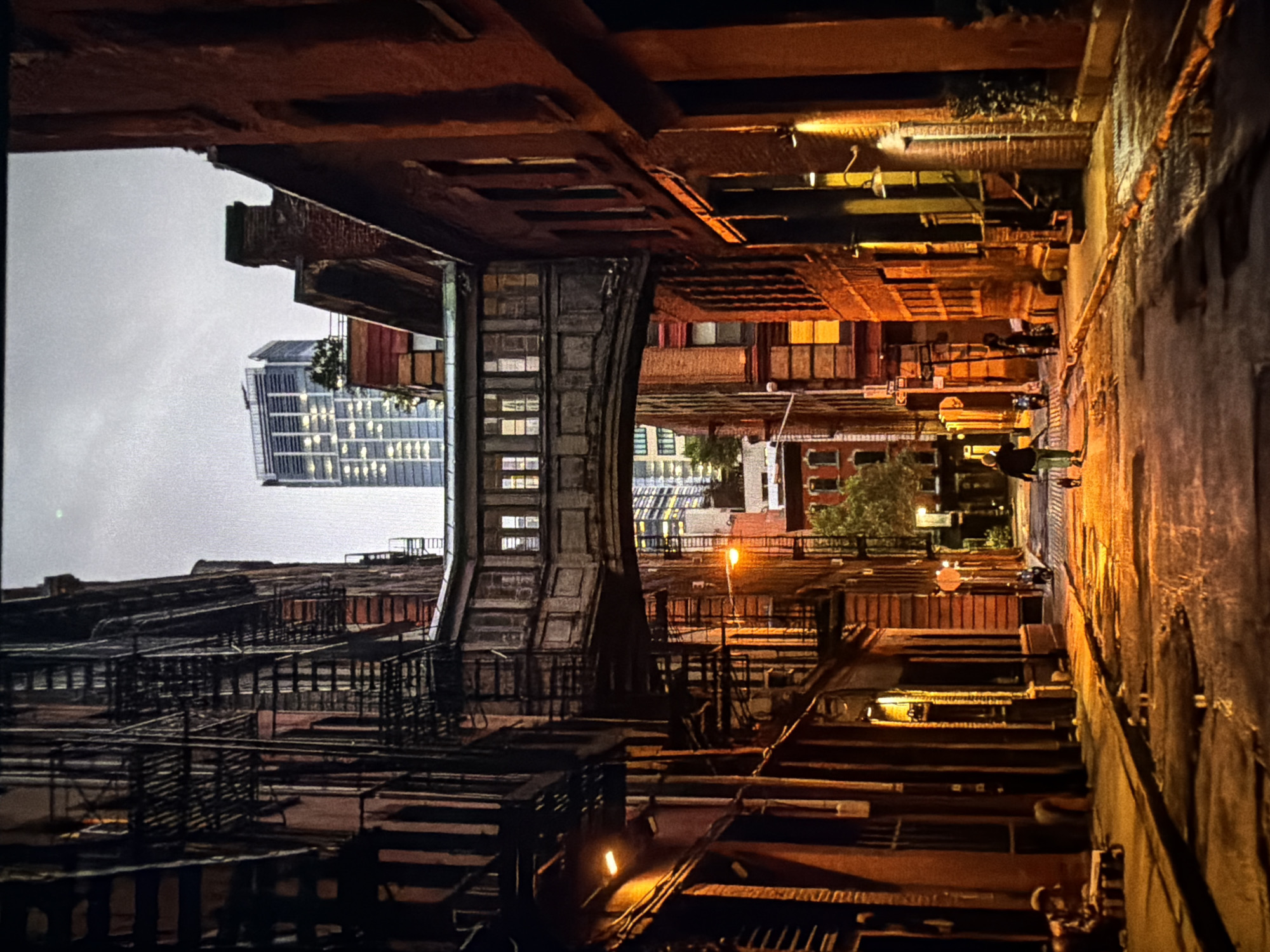 The image displays a narrow, wet urban alleyway or street, viewed from a low perspective, with a pronounced sense of depth. On both sides, multi-story brick buildings with numerous windows and exterior metal fire escapes line the street, exhibiting aged architecture. An enclosed, ornate pedestrian skybridge, constructed of wood or similar material with multiple window panels, spans across the street, connecting the upper levels of the buildings.
The street surface is paved, appears damp and reflective in several areas, suggesting recent rainfall or moisture. Warm, orange-toned artificial light from street lamps and building interiors illuminates the scene, casting reflections on the wet ground and highlighting architectural details. In the immediate foreground, to the left, a person is seated on a low structure. Further into the alley, a solitary individual walks away from the viewer with a small, dark dog on a leash, illuminated by the streetlights. Distant figures and a parked scooter are also visible along the street.
In the far background, contrasting with the historical foreground, a tall, modern skyscraper with numerous brightly lit windows is visible beneath a neutral, overcast sky. The overall ambient light indicates dusk or nighttime. The scene is located in the city of Maidstone, United Kingdom.