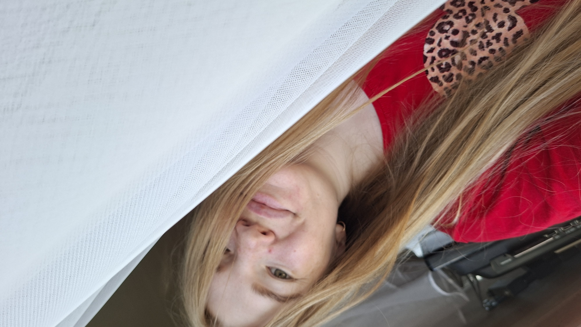 This is a close-up, sideways photo of a young woman in Russia. She has long, blonde hair and is wearing a red top with a leopard print detail on the shoulder. A sheer white fabric, possibly a curtain or veil, is draped across the left side of the frame, obscuring part of her face and hair. Her eyes are open and looking towards the camera. She has a slight smile, and her skin appears fair with some freckles. The lighting suggests it might be daytime. The background is mostly out of focus but appears to be indoors with some dark furniture visible. There is no visible text in the image.