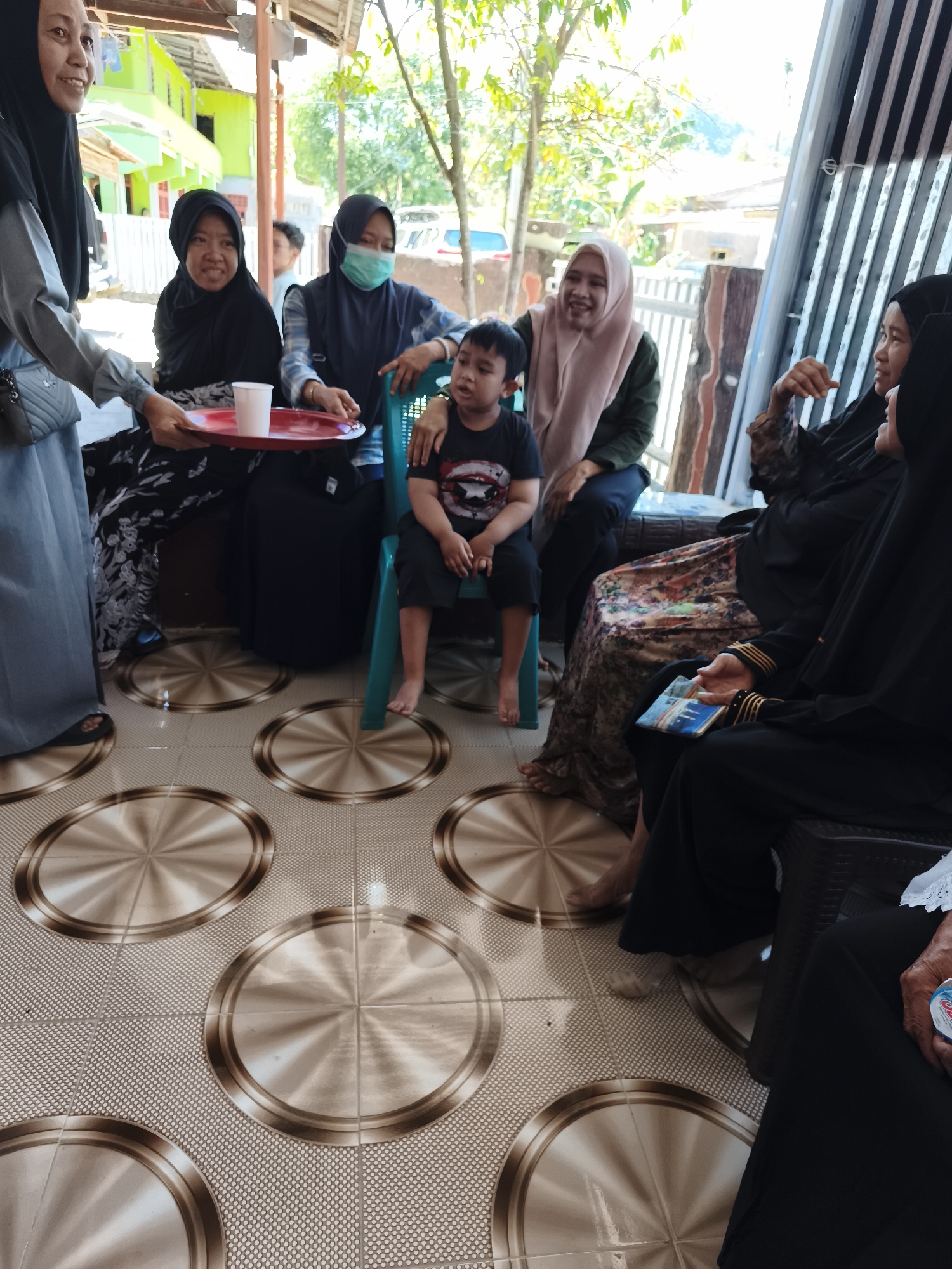 In Tonasa, Indonesia, a group of people are gathered outdoors, possibly for a family event or social gathering. The scene takes place on a tiled patio with a circular pattern. In the foreground, a woman in a grey hijab and a long grey abaya holds a red tray with a white cup. To her right, a woman in a black hijab smiles. Next to her, another woman wearing a blue hijab and a medical mask also appears to be smiling. A young boy in a black t-shirt and shorts sits on a green plastic chair in the center, looking forward. Behind him, a woman in a light pink hijab and green top laughs. To the right, a woman in a black hijab and patterned skirt is seated, holding a small book or card. Further to the right, another woman in a black abaya is visible. The background shows a simple house with bright green walls and a wooden structure. Trees and foliage are present, suggesting a semi-rural or suburban setting. The lighting indicates it is daytime, likely sunny.