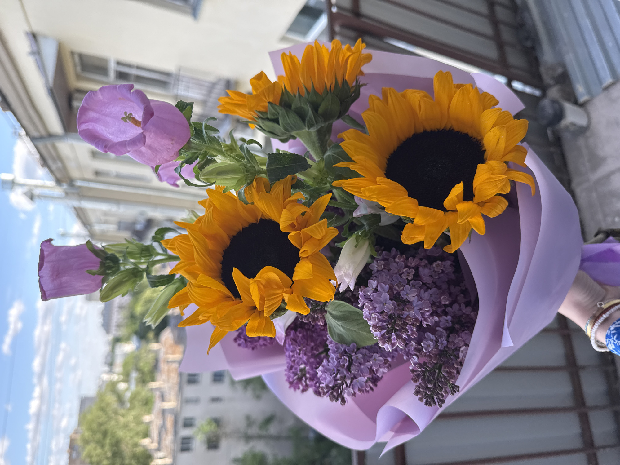 A bouquet of flowers, including sunflowers, lilac, and campanula, is held in front of a city background. The bouquet is wrapped in lavender-colored paper. The sunflowers have dark centers and bright yellow petals. The lilac is purple and clustered. The campanula are light purple, bell-shaped flowers. The background shows buildings in Moscow, Russia under a blue sky. A hand holding the bouquet is visible in the lower right corner, with bracelets on the wrist.