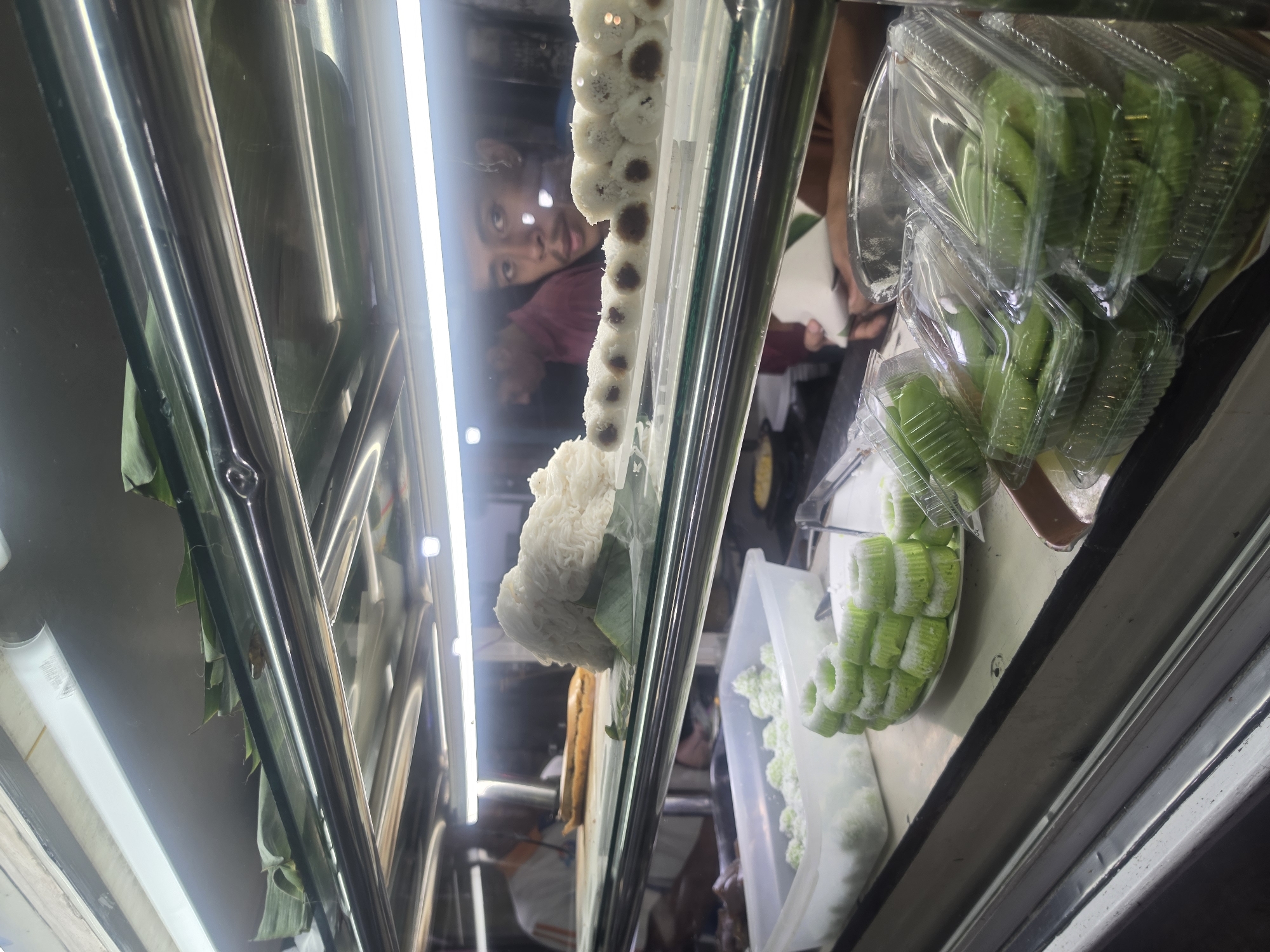 This is a photograph taken inside a food display case, likely in a market or restaurant in the city of Medan, Indonesia. The display case has multiple shelves, illuminated by fluorescent lights. On the top shelf, a pile of white, stringy food, possibly a type of noodle or sweet confection, sits on a green leaf. Next to it, a row of small, white, cylindrical pastries with dark centers are arranged on a white plate.
On the shelf below, a white plastic container is filled with small, pale green, ball-shaped sweets dusted with white powder. In front of this container, a stack of green, ring-shaped cakes, also dusted with white, are arranged on a white plate. To the right of these cakes, several clear plastic containers are stacked, each filled with green, flat, disc-shaped pastries.
In the background, two individuals are visible. One, wearing a white shirt with orange stripes, appears to be standing behind the display. Another, a young man with dark hair, is looking directly at the camera through the glass of the display case. The overall environment suggests a commercial food vendor.