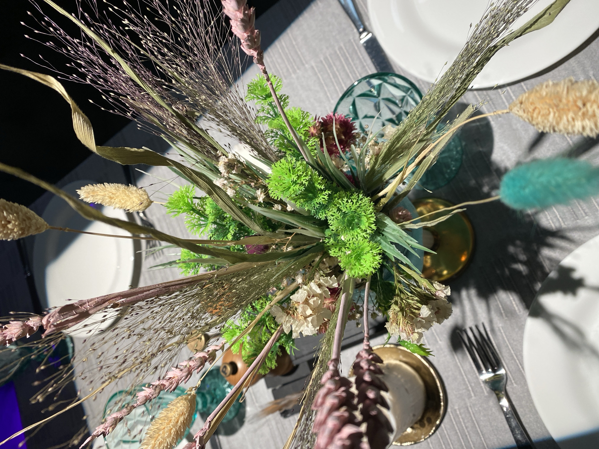 The image displays a close-up, high-angle view of a table setting, likely for a meal. The table is covered with a grey tablecloth. Dominating the center is a floral arrangement of dried grasses and greenery in a gold vase. Scattered around the arrangement are white plates, a silver fork, and a green glass. The lighting suggests it might be indoors with a strong light source creating shadows on the table. No people are visible in the image, and no specific location in Myl'tsi, Ukraine, can be identified.