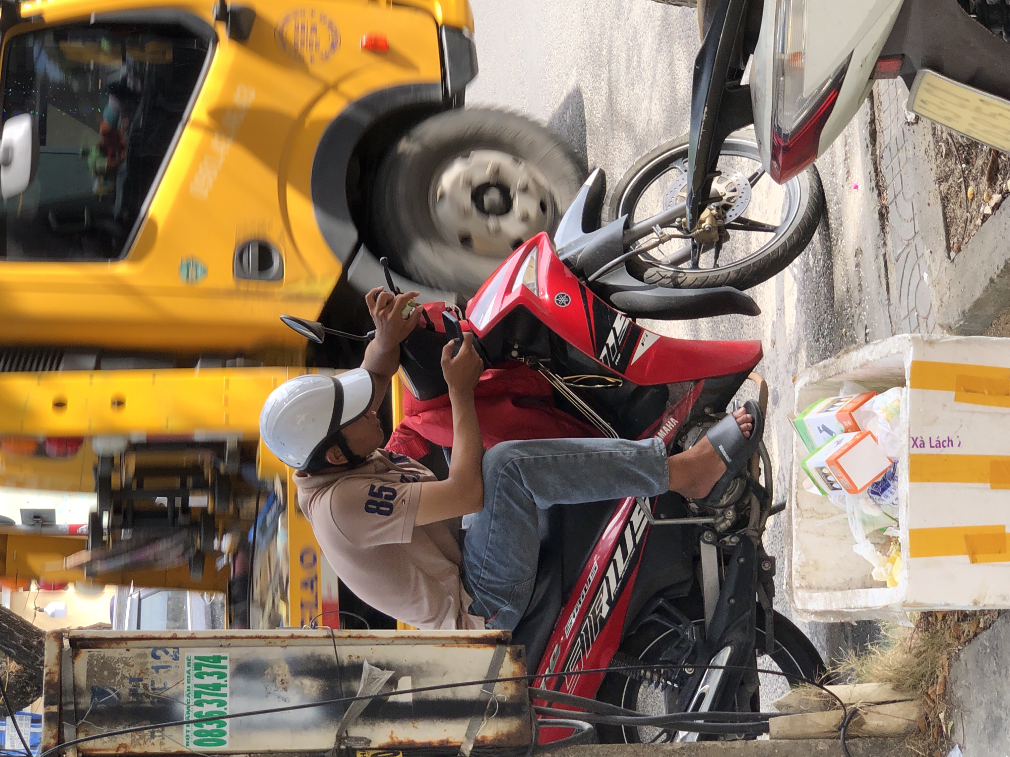A Vietnamese man wearing a white helmet and a brown polo shirt with the number "85" on it is sitting on a red and black motorcycle. He is looking down at his phone, which he is holding in his right hand. His left hand is on the handlebars. The motorcycle is parked on the side of a street in Da Nang, Vietnam. In the background, a yellow truck is driving by. To the left of the motorcycle, there is a utility pole with a sign that has Vietnamese writing and a phone number on it. In the foreground, there is a white styrofoam box filled with trash. The ground is paved with asphalt and some cobblestones. The lighting suggests it is daytime, likely sunny.