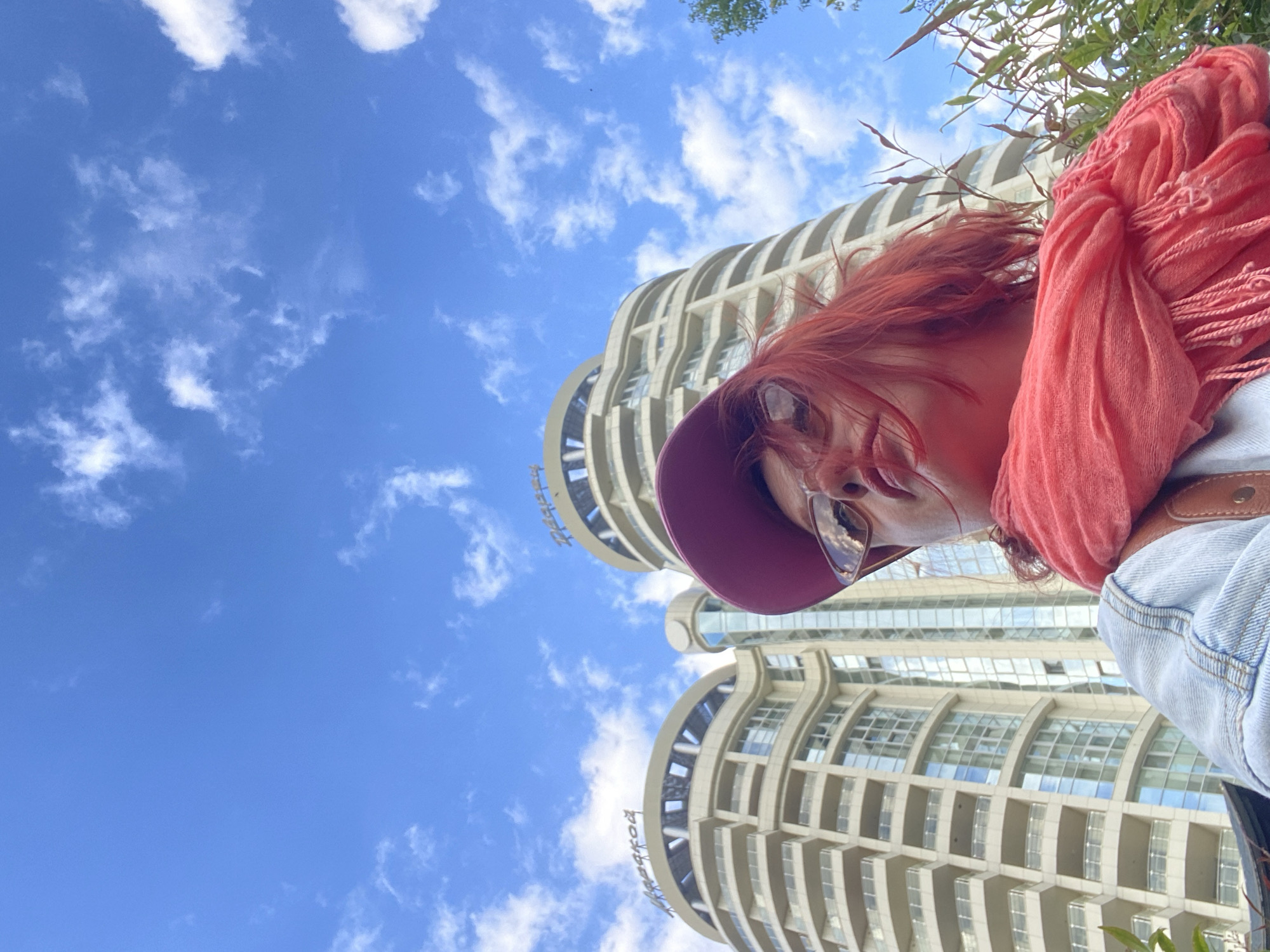 A low-angle, outdoor shot shows a person, presumably female, wearing a purple hat, sunglasses, and a pink scarf, positioned against a backdrop of a blue sky with scattered white clouds. In the background, a tall, curved building with numerous windows is visible. The image is taken from below, giving the impression that the person is looking down at the camera. This building can be identified as part of the Sochi City business center in Sochi, Russia.