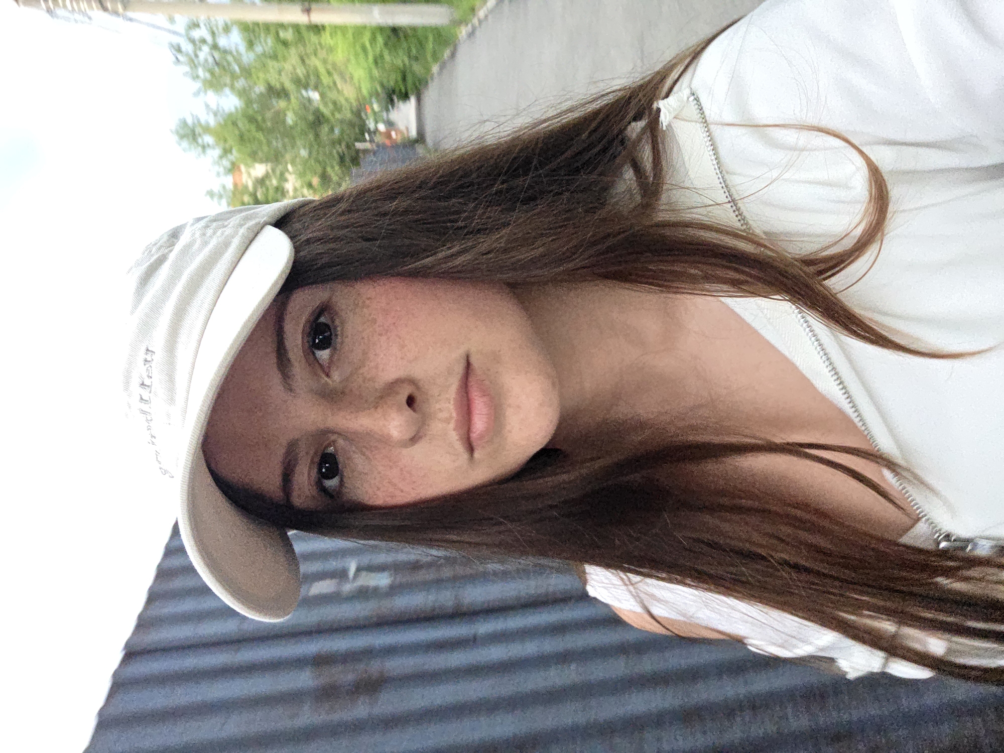 A young woman with long, brown hair and freckles across her nose and cheeks is captured in a selfie. She is wearing a white baseball cap and a white zip-up jacket. Her hair is partially covering her face, framing her eyes and lips. The image is taken outdoors, with a blurred background suggesting a street or sidewalk with trees and a grey, corrugated surface in the foreground. The lighting suggests it is daytime, possibly overcast given the diffused light. The woman's expression is neutral, with her gaze directed towards the camera. The image is oriented sideways, with the woman appearing to be looking up. There is some faint text visible on the brim of her cap, but it is illegible.