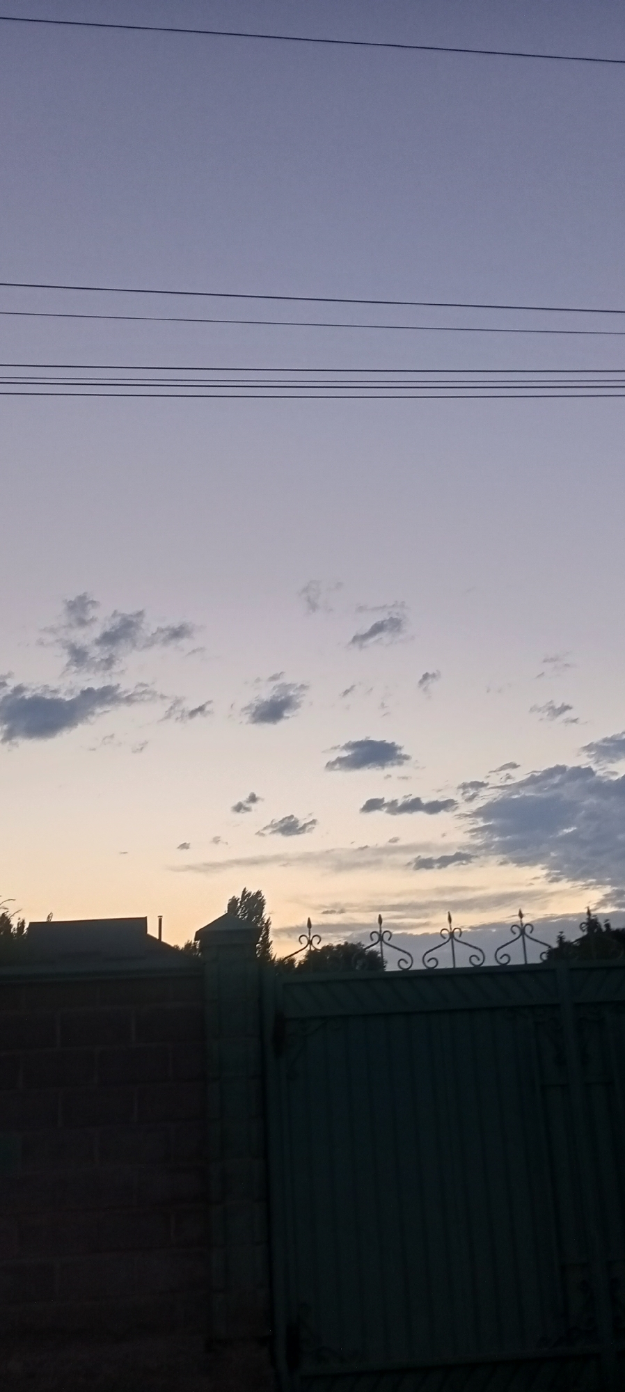 The image depicts a twilight scene over a residential area in Romanovka, Kyrgyzstan. A soft, lavender sky dominates the upper portion of the frame, with several thin, dark horizontal lines, likely power lines, stretching across it. Below the sky, a scattering of small, wispy clouds are visible. The foreground is partially obscured by a dark, brick wall on the left and a dark green, corrugated metal gate with decorative wrought-iron elements on the right. The top edge of the gate features a row of ornate finials. A sliver of a dark-roofed building is visible behind the brick wall, and trees can be seen in the distance beyond the gate, silhouetted against the lighter sky. The overall impression is one of a quiet, suburban evening.
