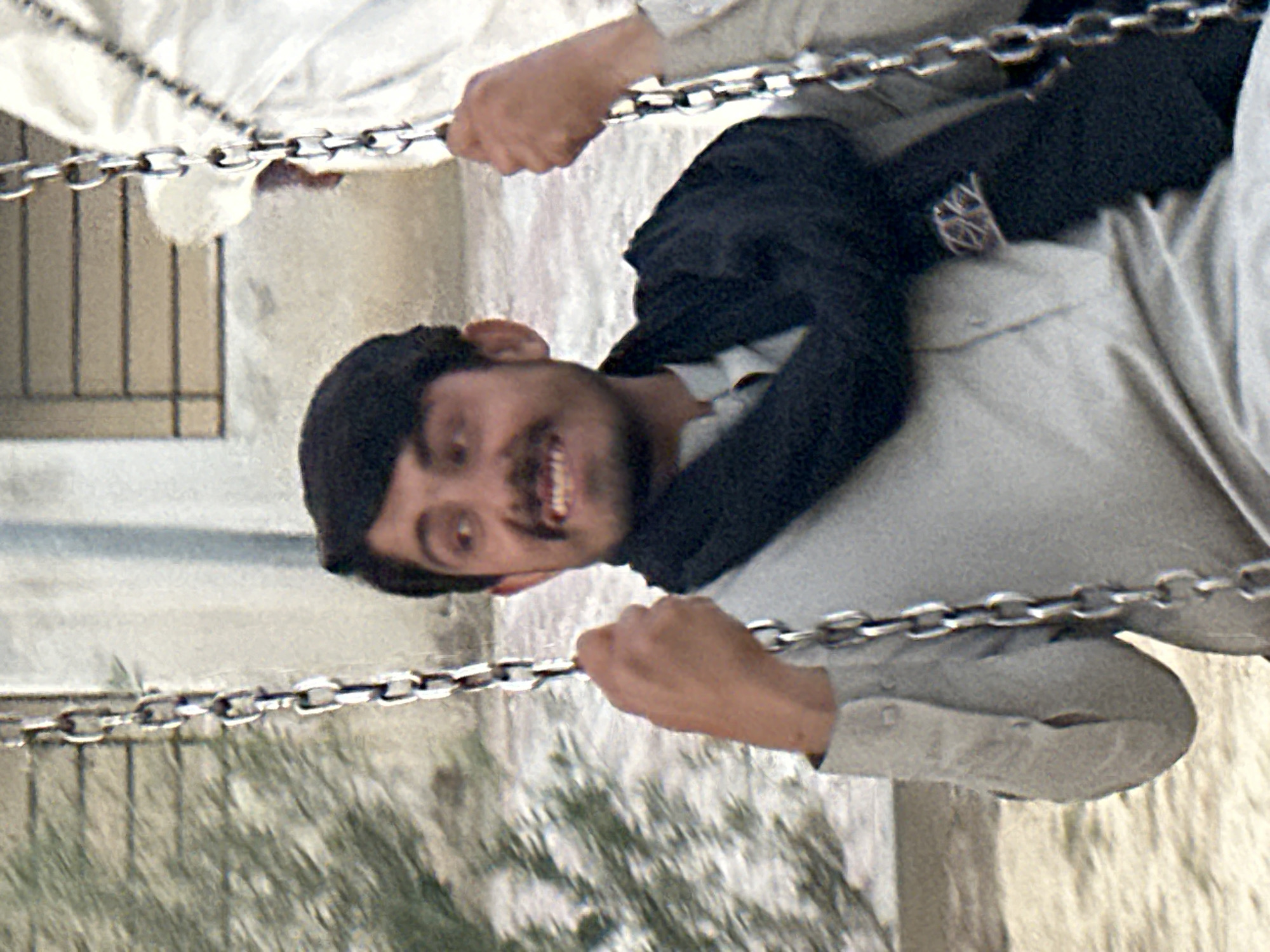 The rotated image captures a person, likely an adult male, holding onto two parallel metal chains, appearing to be upside down. He is wearing a light-colored long shirt (possibly a shalwar kameez) and a dark jacket or shawl. His face is visible, featuring a mustache and a wide, joyful smile, with his teeth showing. His eyes are open, looking directly towards the viewer. Both hands are firmly gripping the chains, one hand near the top-left of the frame and the other near the bottom-right, relative to his body.
In the background, there are light-colored structures, possibly walls or buildings, and blurred green foliage. A window with vertical bars is visible in the upper-left quadrant. The overall lighting suggests it is daytime. The man's attire and features hint at a South Asian or Middle Eastern context. The activity appears to be an acrobatic feat, a game, or a performance, given his upside-down position and evident enthusiasm. No discernible text is visible in the image.