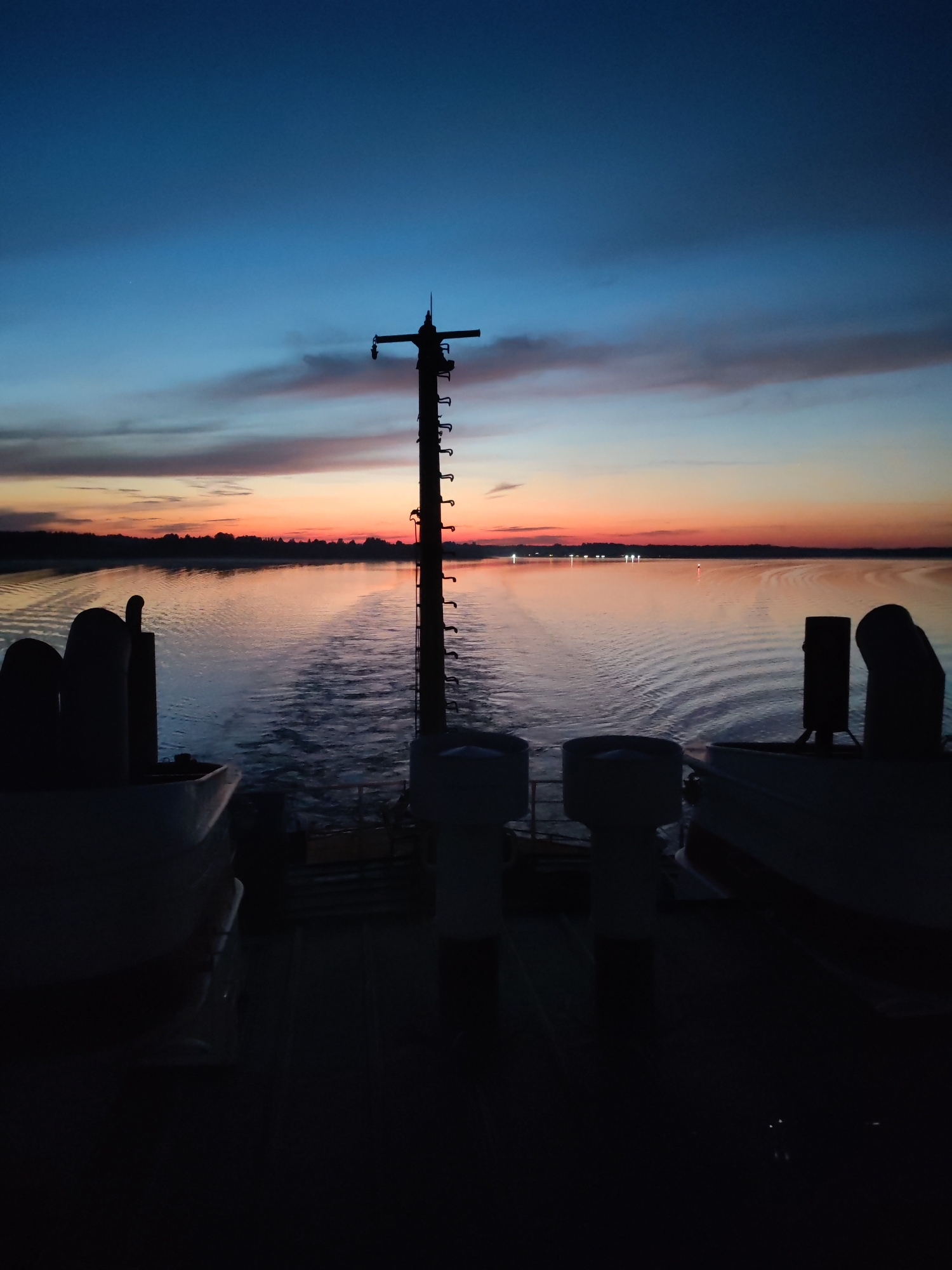The image captures a sunset or sunrise scene from the deck of a vessel. The sky displays a gradient of deep blue at the top, transitioning through lighter blues and streaks of pink and orange towards the horizon. A tall, dark pole with what appear to be lights or equipment is silhouetted in the center of the frame, extending from the deck upwards. The water in front of the vessel shows a wake, indicating movement. In the foreground, the dark silhouettes of parts of the vessel are visible, including curved structures that could be railings or equipment. The distant shoreline is lined with trees and dotted with faint lights. The overall impression is one of tranquility and the vastness of the open water.