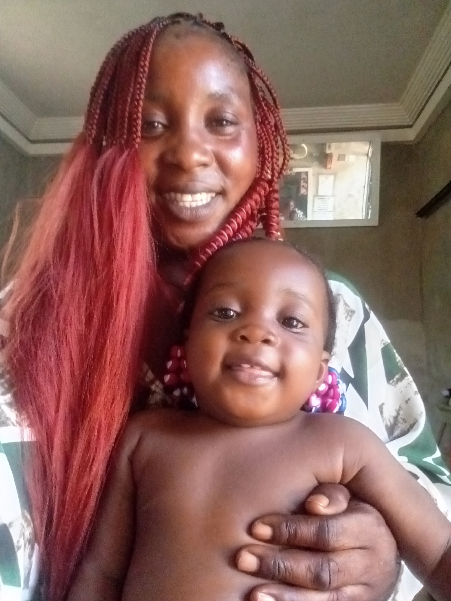 This indoor portrait features a woman holding a baby. The woman, with dark skin, is positioned in the background, her face visible in the upper part of the frame. She has long, red braided hair, with some braids styled and other strands flowing loosely down her left side. She is wearing a patterned garment with white, green, and black designs. The woman is smiling broadly, showing her teeth, and her eyes are looking directly at the viewer.
In front of her, held securely, is a baby with dark skin. The baby is shirtless, revealing bare shoulders and chest, and appears to be wearing a necklace or beaded accessory with pink, purple, and white beads around its neck. The baby is also smiling gently, looking towards the camera, with some early teeth or gums visible. The woman's hands are visible, supporting the baby around its midsection.
The setting is an indoor room, likely a home. A light-colored wall forms the background, with a mirror or framed picture visible behind the woman's right shoulder, reflecting indistinct shapes that resemble papers or other items. The ceiling features decorative molding. The lighting suggests it is daytime. Both the woman and the baby are smiling, conveying a sense of happiness and affection. No discernible text is visible in the image.