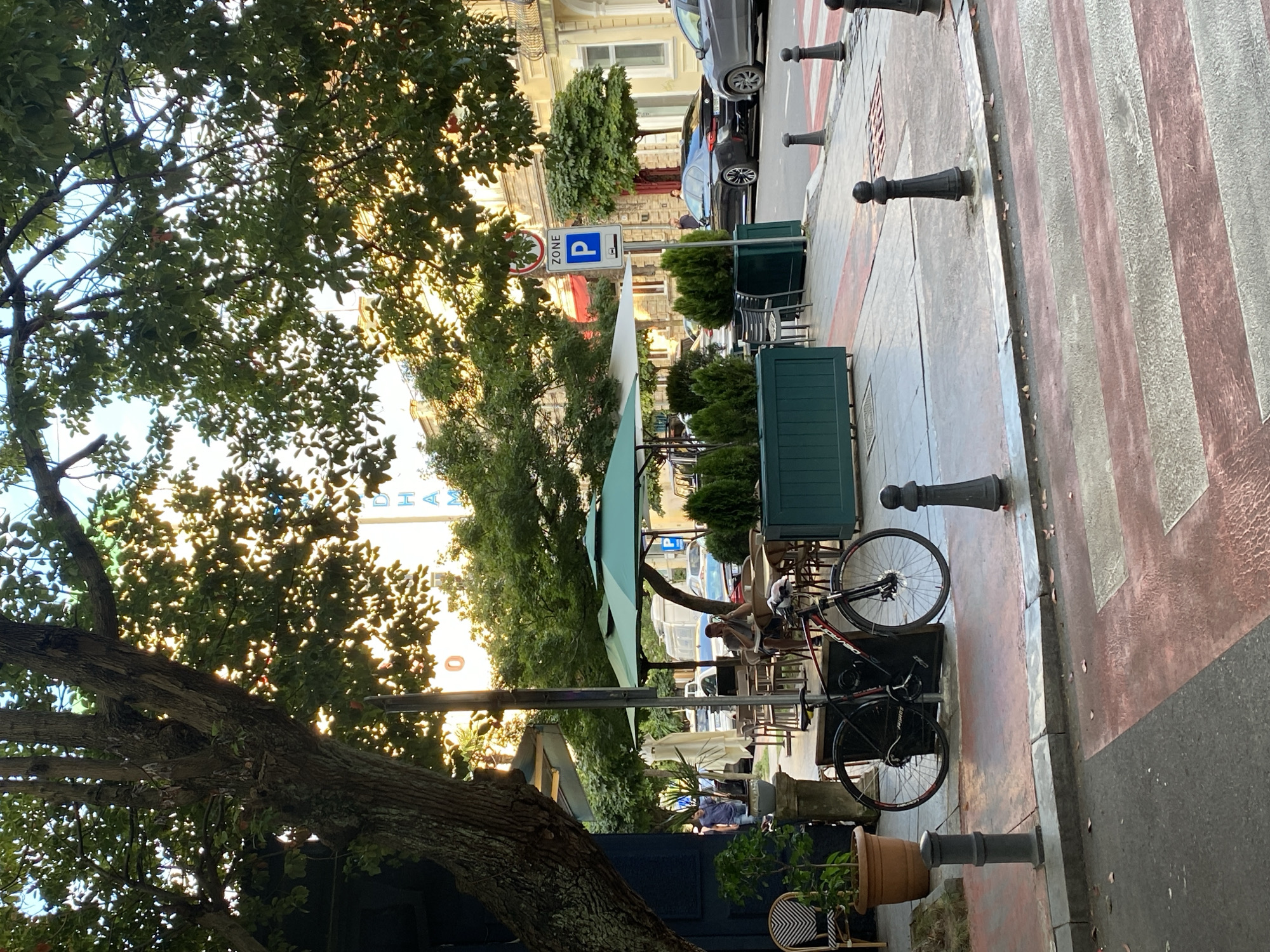The image captures a street scene in Batumi, Georgia, with a focus on an outdoor cafe area situated on a sidewalk next to a street. The sun is shining, casting dappled light through the leaves of a large tree in the foreground. A black bicycle is parked next to a dark trash receptacle in front of the cafe. The cafe features green umbrellas providing shade over tables and chairs. A person is seated at one of the tables, facing away from the camera. Several other tables and chairs are visible. Beyond the cafe, the street is lined with buildings and trees. A parking zone sign with a "P" symbol is posted on a pole. Cars are parked and driving on the street. A pedestrian crossing with red and white stripes is visible in the lower part of the image. The overall impression is a quiet, daytime scene in an urban environment.