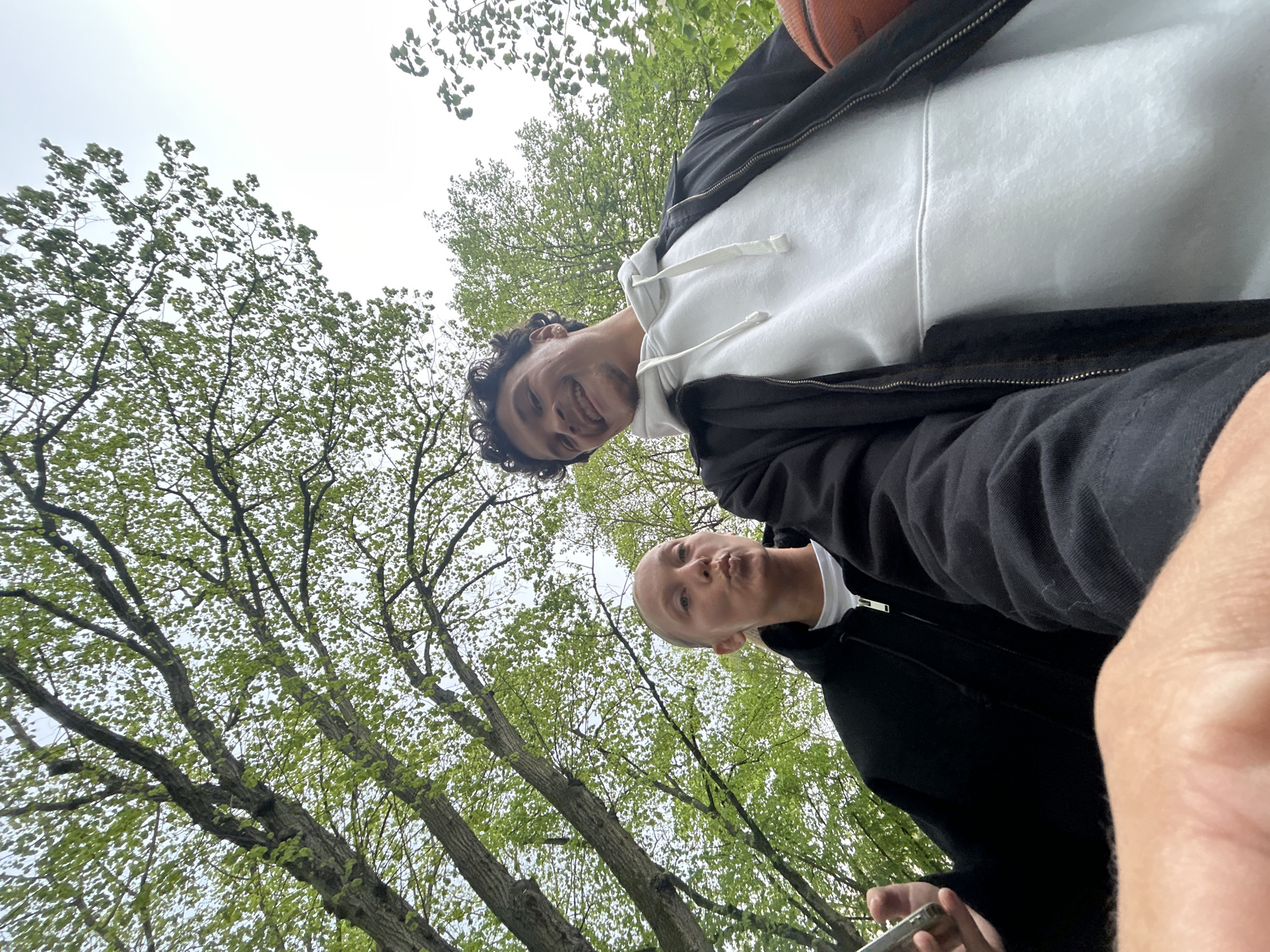 Two people are looking down at the camera from a low-angle perspective, likely in an outdoor park setting. The person at the top of the frame is smiling and wearing a white hooded sweatshirt with a black zip-up jacket. They are holding a basketball partially visible in the top right corner. The person below is making a kissing face and wearing a white t-shirt with a black zip-up jacket. They are holding a smartphone in their hand, presumably taking the picture. The background consists of trees with green foliage. The photo was taken during the day, under an overcast sky. It's difficult to be certain of the location in Noisiel, France, but the style of the trees suggests it could be a park.