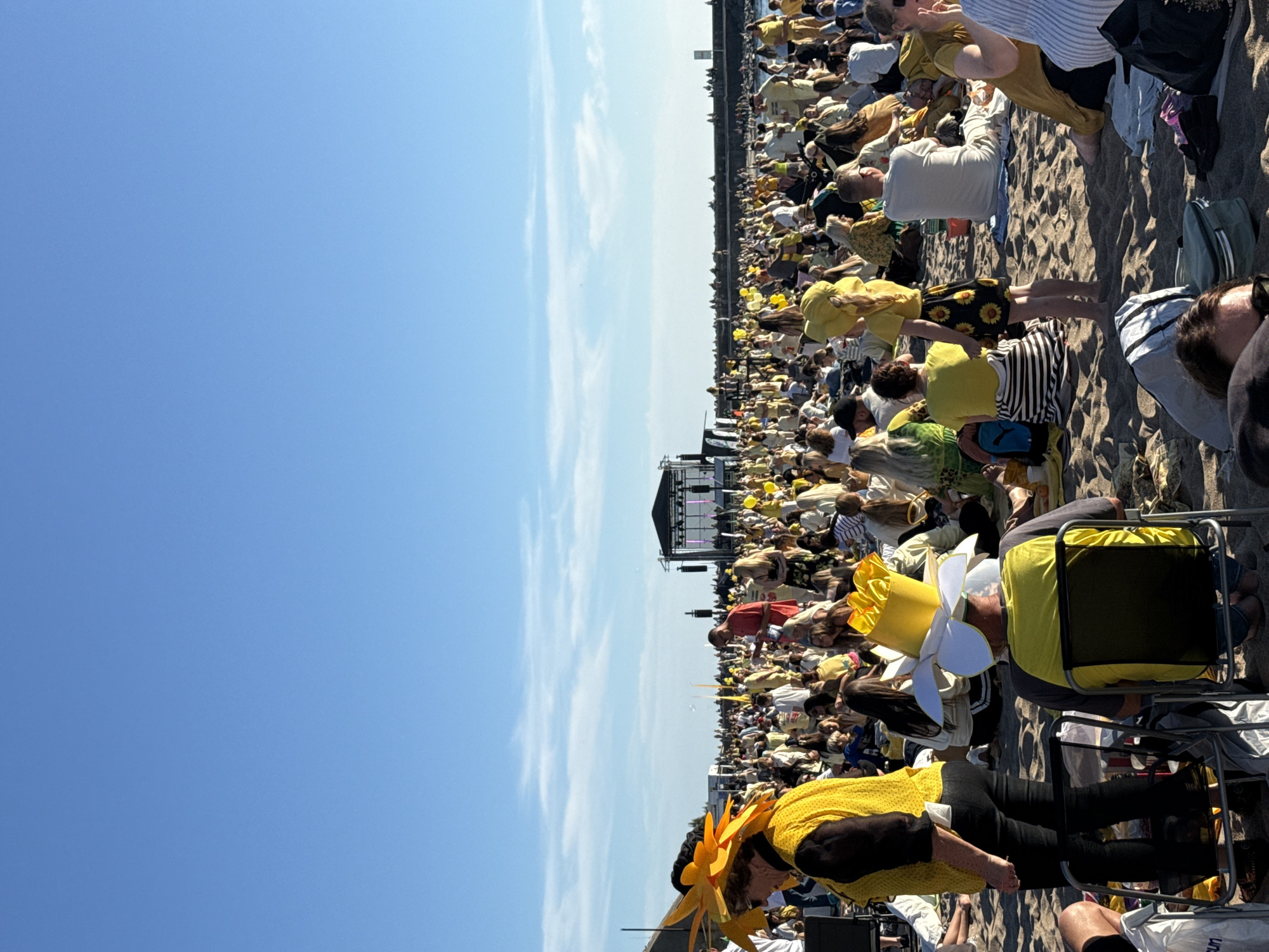 A large crowd of people are gathered on a sandy beach, facing a stage with a black tent-like structure and lights. Many individuals are wearing yellow clothing, and some have elaborate yellow hats, including one shaped like a flower and another resembling a sunflower. The sky is clear and blue with some wispy clouds. The overall scene suggests an outdoor event or festival, possibly a concert or performance, taking place on a sunny day. The people are sitting, standing, and milling about on the sand, with a stage set up in the distance.