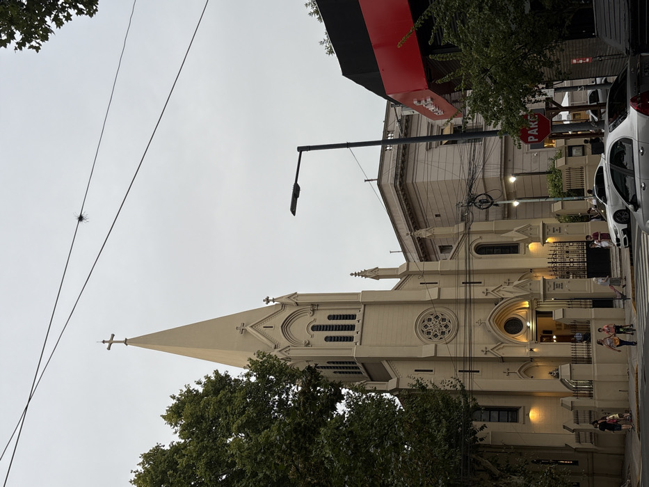 A tall, light-colored church with a pointed spire dominates the scene. The church features arched windows and a decorative rose window. A green tree obscures part of the lower section of the church. Above, power lines run diagonally across the sky. A street lamp extends out from the building next to the church. People are visible on the sidewalk near the church entrance. A "Pare" (Stop) sign and a white car are located on the right side of the image. The sky is overcast.