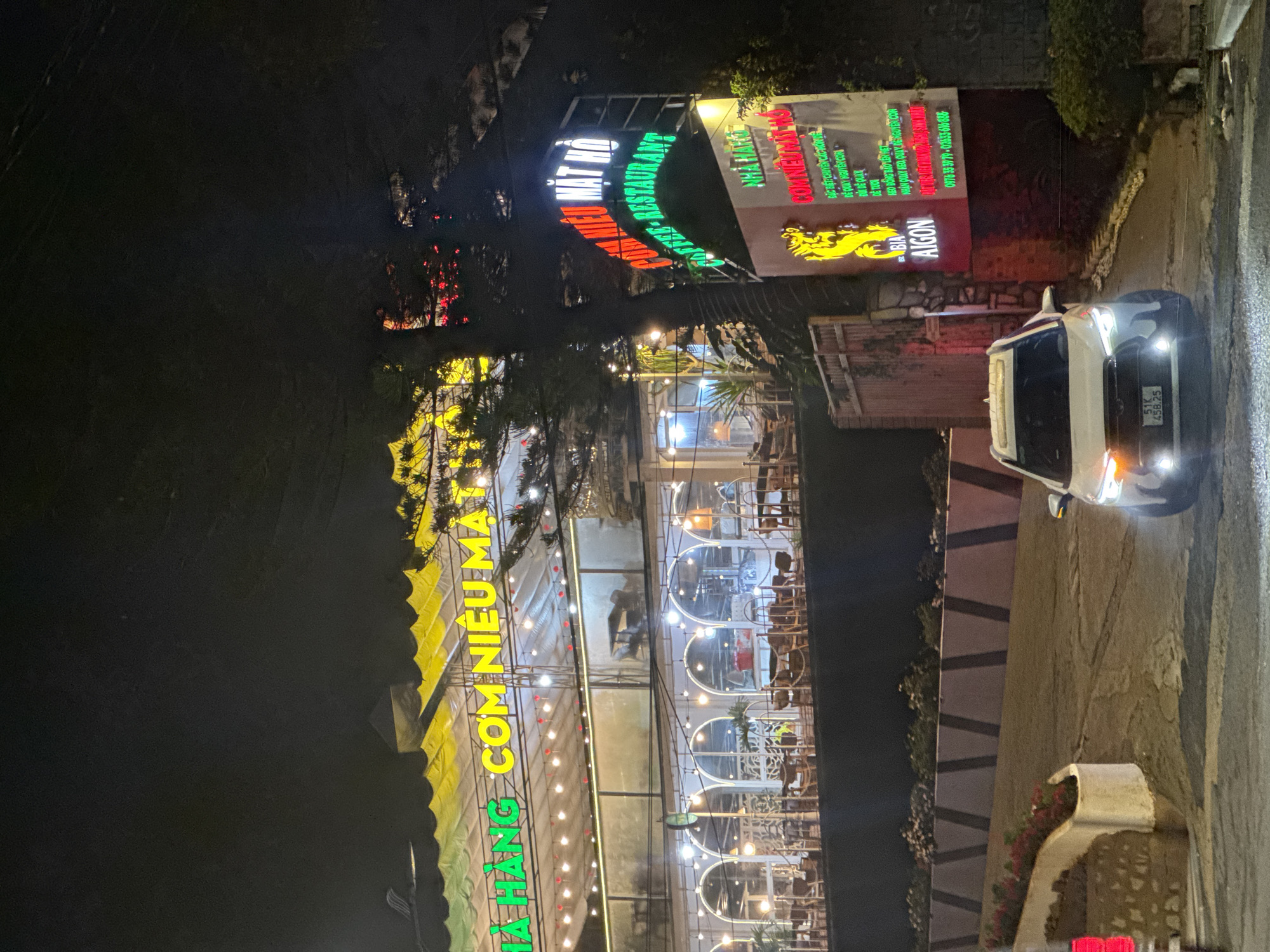 The image depicts the front of the "HA HANG COM NIEU MA" restaurant at night. The restaurant's name is prominently displayed in large, illuminated yellow and green letters. A white car is parked on the side of the road in front of the restaurant. The car's license plate reads "51K 458.25." Another sign for the restaurant is visible, including a dragon. The sign advertises Com Nieu and features phone numbers. The street appears to be wet, and the background is dark. The restaurant appears to be located in Da Lat, Vietnam.