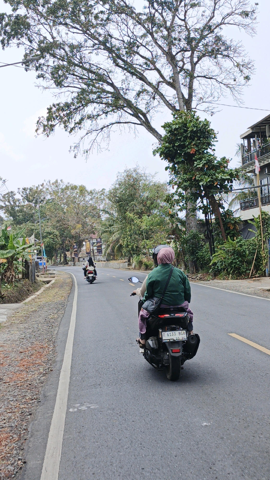 The image captures a view from behind two individuals riding on a motorcycle on a paved road in Sukaresik, Indonesia. The person in the foreground, wearing a green top and a pink headscarf, is seated behind the driver. They are carrying a black bag on their shoulder and another bag attached to the back of the motorcycle. The motorcycle's license plate reads "R 6133 BGB". In the background, another motorcycle with two passengers is visible further down the road. The environment features lush green trees and vegetation lining the road, with a building visible on the right side of the frame. The sky is overcast. The road has a white line marking the lane and a yellow line on the right. The area to the left of the road appears to be unpaved and covered with gravel and dried leaves.