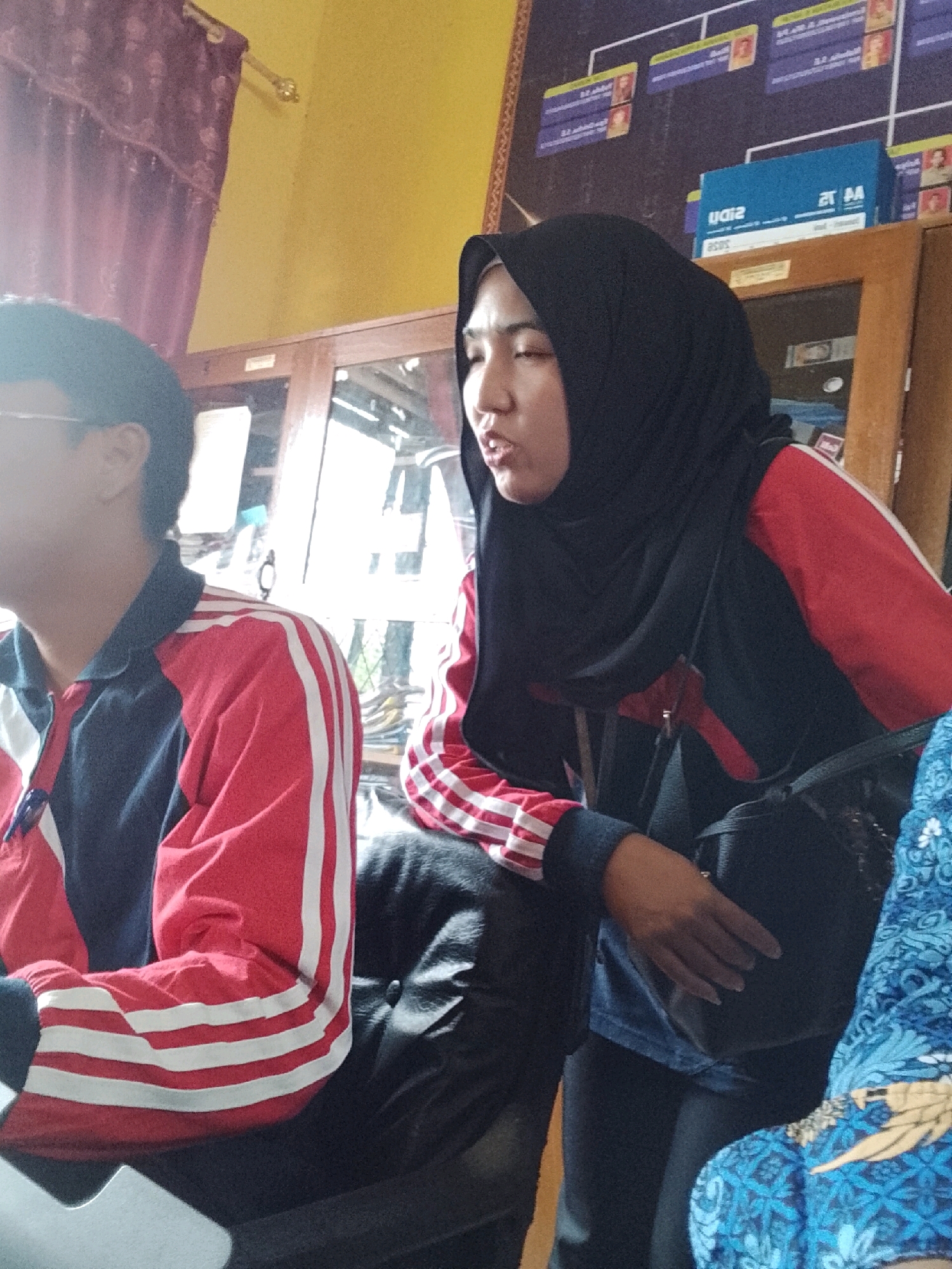 The image captures two individuals in what appears to be an indoor setting, possibly a classroom or office, in Jepara, Indonesia. On the left, a person wearing glasses and a dark shirt with red and white stripes on the sleeves is partially visible. The person on the right is wearing a black hijab and a red and dark jacket with white stripes on the sleeves, and appears to be speaking. A black handbag is slung over their shoulder. The background reveals wooden furniture, including what looks like a cabinet or shelf with items on it, and a bulletin board with charts and pictures. The lighting suggests it is daytime.