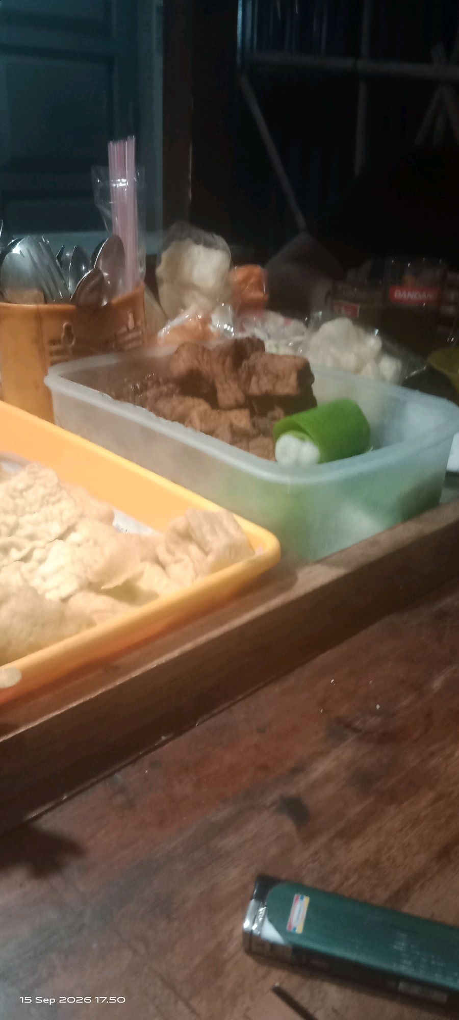 A close-up, slightly blurry, eye-level shot captures a table setting, likely in Merbung, Indonesia, given the timestamp. The foreground is dominated by a dark, wooden surface with a green and silver cigarette lighter lying horizontally on the right side. To the left of the lighter, a faint shadow suggests a seated person. The mid-ground features two containers of food. The container on the left, a yellow oblong dish, holds what appear to be fried snacks, possibly crackers or chips. To its right, a clear plastic rectangular container holds pieces of fried food, possibly tempeh, and a single green item, possibly wrapped tofu or a vegetable, rests on top. In the background, out of focus, are more containers and what looks like a bottle cap. The overall lighting is dim, contributing to the blurred appearance of the scene.