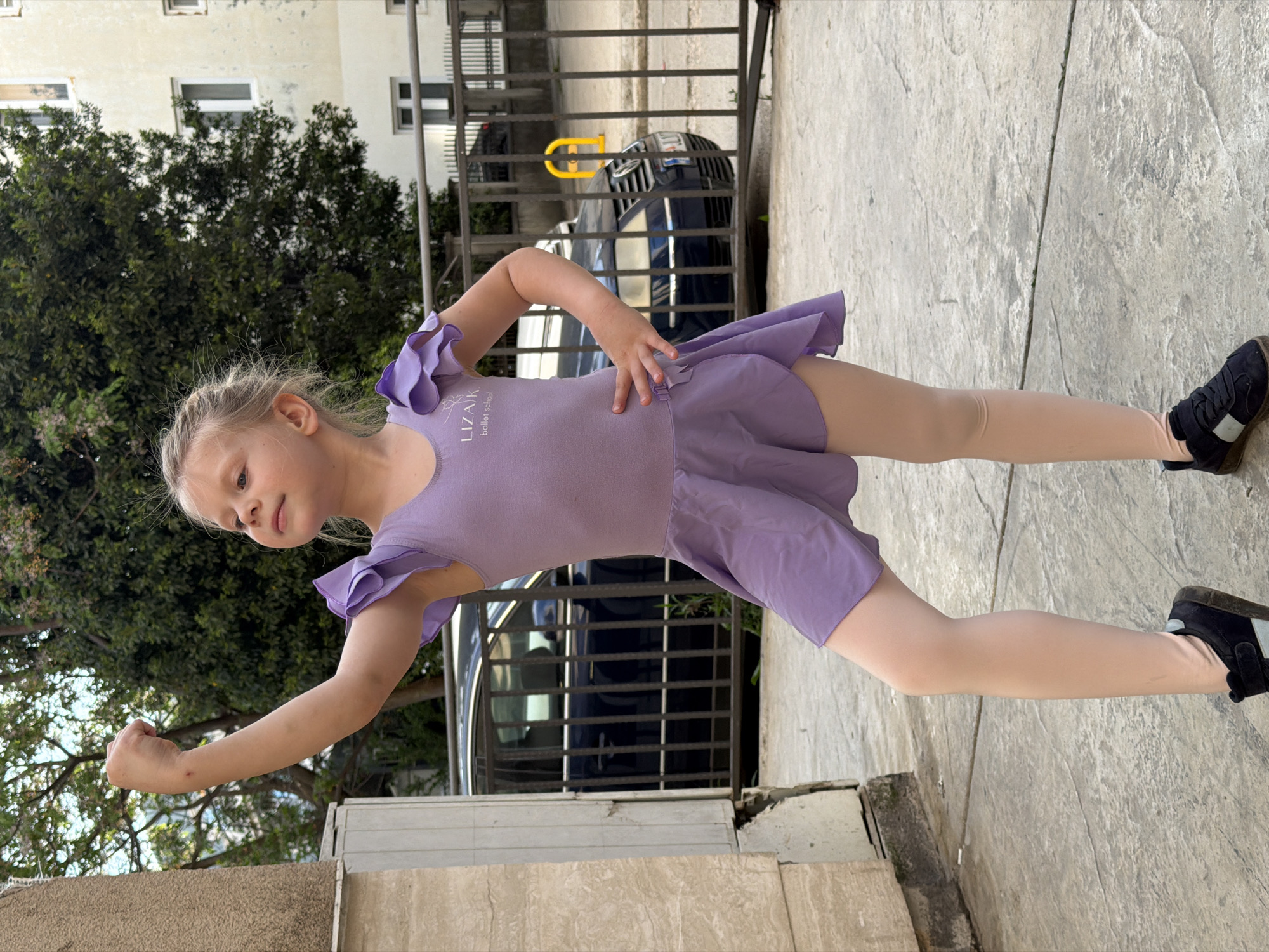 A young, blonde-haired girl, appearing to be around preschool or early elementary school age, is the central subject of this vertically oriented outdoor photograph. She is wearing a light purple ballet leotard with ruffled cap sleeves and an attached skirt, light-colored tights, and dark sneakers with white soles and straps. Her leotard has text "LIZA.K ballet school" visible on the chest.
The girl stands with her right arm bent at the elbow, hand on her hip, and her left arm extended forward and slightly upward, with her hand clenched into a fist. She looks towards the left of the frame with a focused, pleasant expression.
The setting appears to be an urban or suburban outdoor area in Budva, Montenegro. The ground beneath her is paved with light-colored, textured concrete. In the background, behind a dark metal railing, a dark-colored car is partially visible. Further back, there's a light-colored building with multiple windows, and lush green trees are prominent, suggesting a warm climate. The bright, even lighting indicates it is daytime with clear weather.