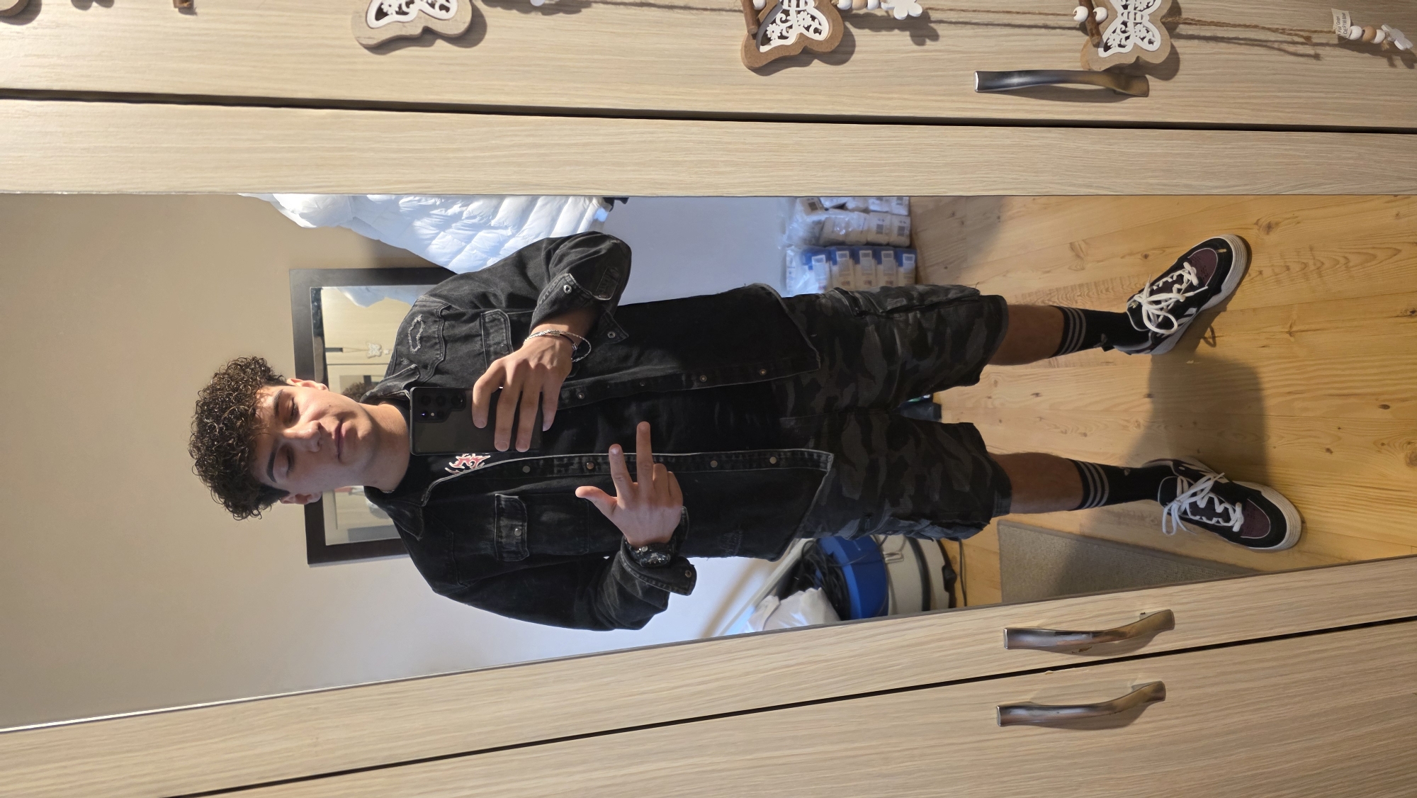 A young male is taking a mirror selfie indoors. He stands on a wooden floor wearing camo shorts, black socks with white stripes, black and white sneakers, a black t-shirt, and a black jean jacket. He holds a smartphone in one hand, making a peace sign with the other. The reflection shows parts of a bed with white bedding, storage containers, and a blue object. The mirror is mounted on a light wood-grain cabinet with two silver handles. Decorative wooden ornaments hang above the mirror. The location is not identifiable as being in Dobrich, Bulgaria, based on the image.