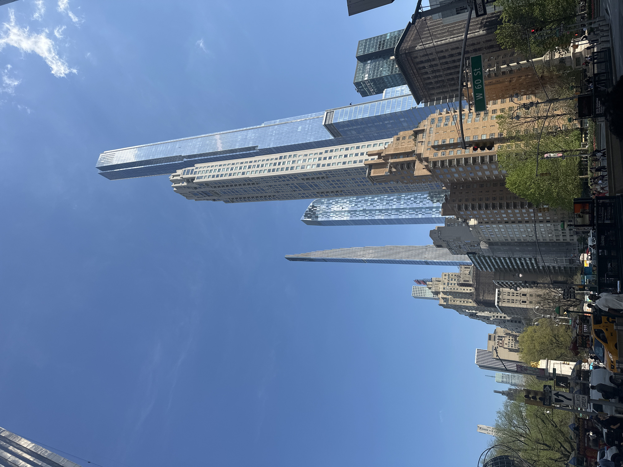 The image captures a street scene in New York City, USA, characterized by towering skyscrapers against a clear blue sky. The buildings exhibit a range of architectural styles, from modern glass structures to older, more ornate designs. A street sign indicates the location as W 60 St. Below, a bustling street with pedestrians, vehicles including yellow taxis, and typical urban infrastructure such as traffic lights and street signs are visible. The presence of trees suggests a park or green space nearby.