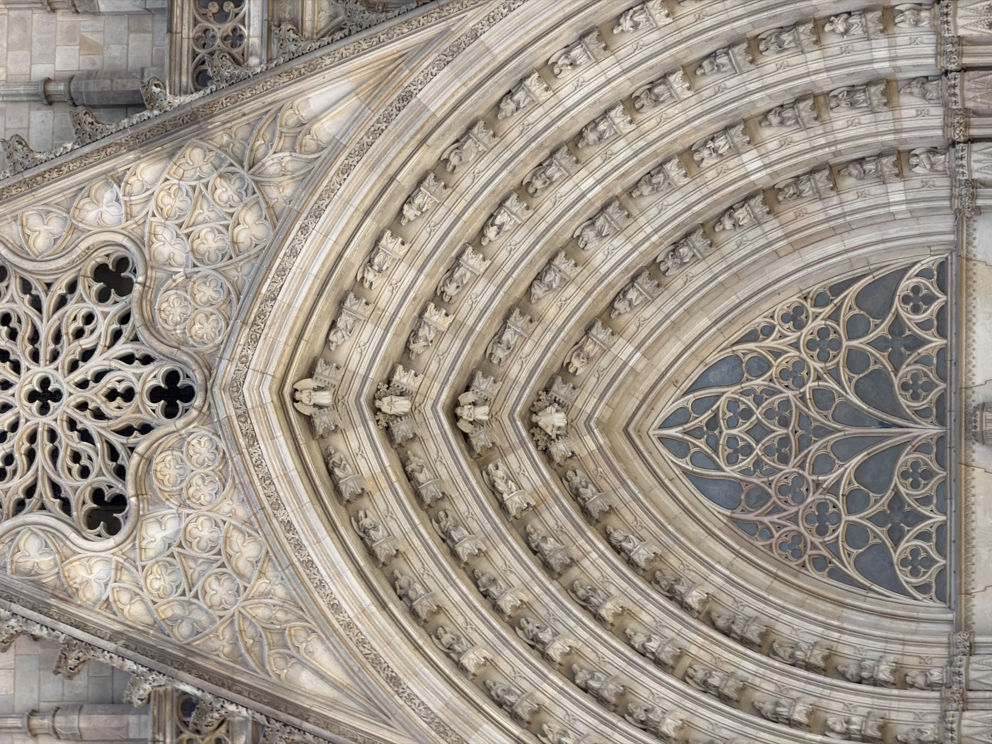 The image shows a detailed view of the facade of the Barcelona Cathedral, a Gothic cathedral located in Barcelona, Spain. The photograph focuses on the upper part of the main entrance archway, which is elaborately decorated with intricate stonework. The arch is segmented into multiple layers, each adorned with small sculptures of figures, possibly angels or saints, in niches. These figures are densely packed, creating a repetitive pattern along the curved surfaces of the arch. Above the main arch, there is a large, ornate rose window with complex geometric tracery, also rendered in stone. The entire facade is constructed from light-colored stone, showing signs of age and weathering. The architectural style is distinctly Gothic, characterized by pointed arches, detailed carvings, and a sense of verticality.
