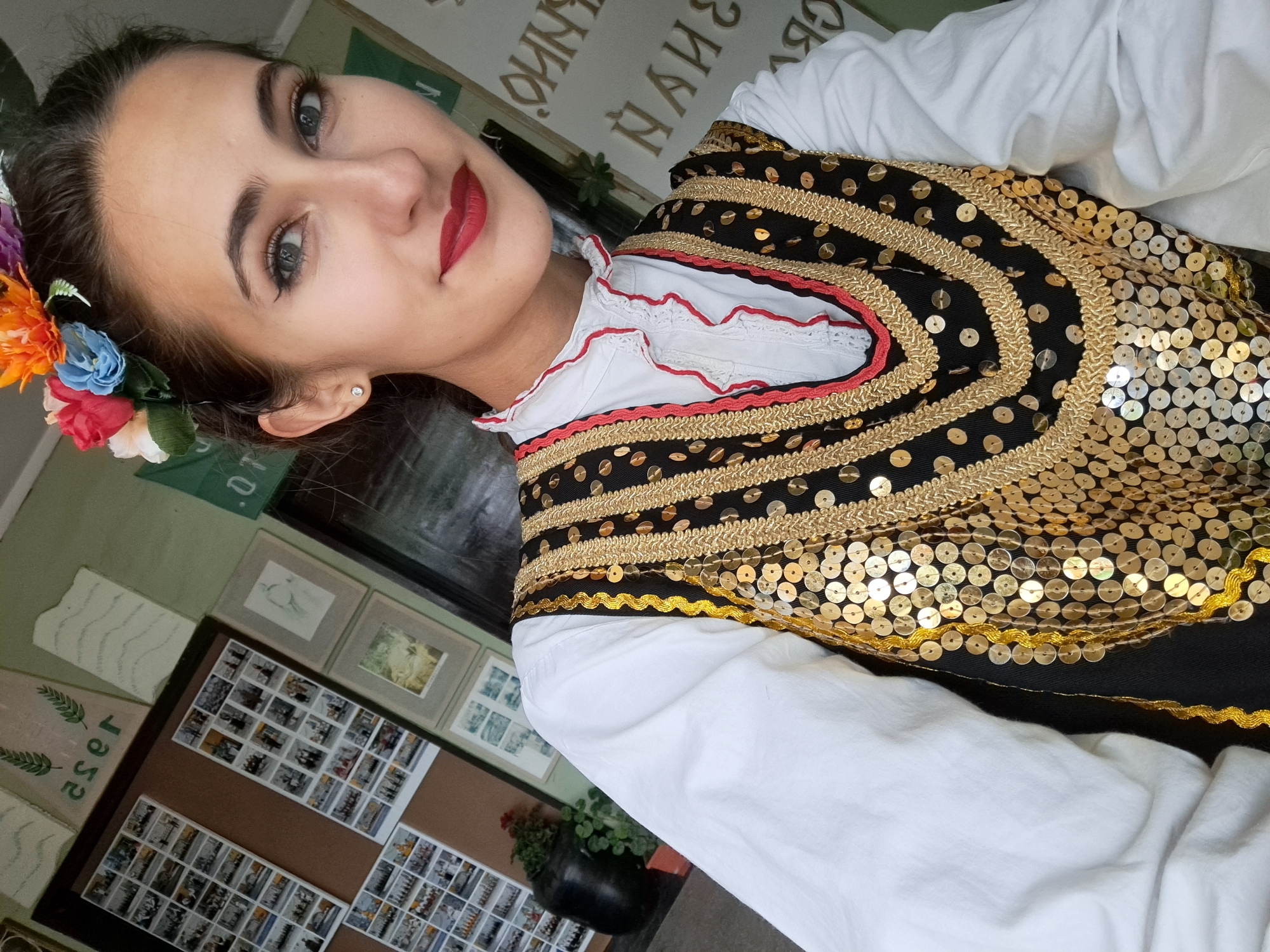 A woman is posing for a selfie in what appears to be an indoor environment. She is wearing a traditional Bulgarian folk costume, consisting of a white blouse with red trim and a heavily embellished black vest covered in gold sequins and trim. She has a floral wreath in her hair.
Behind the woman, there is a wall with various posters and framed images displayed on it. A potted plant sits on a table near the wall. The environment suggests a cultural or historical setting, perhaps related to Bulgarian heritage. While the location in the city of Dragoman, Bulgaria, is specified, it cannot be verified solely from the image's content.