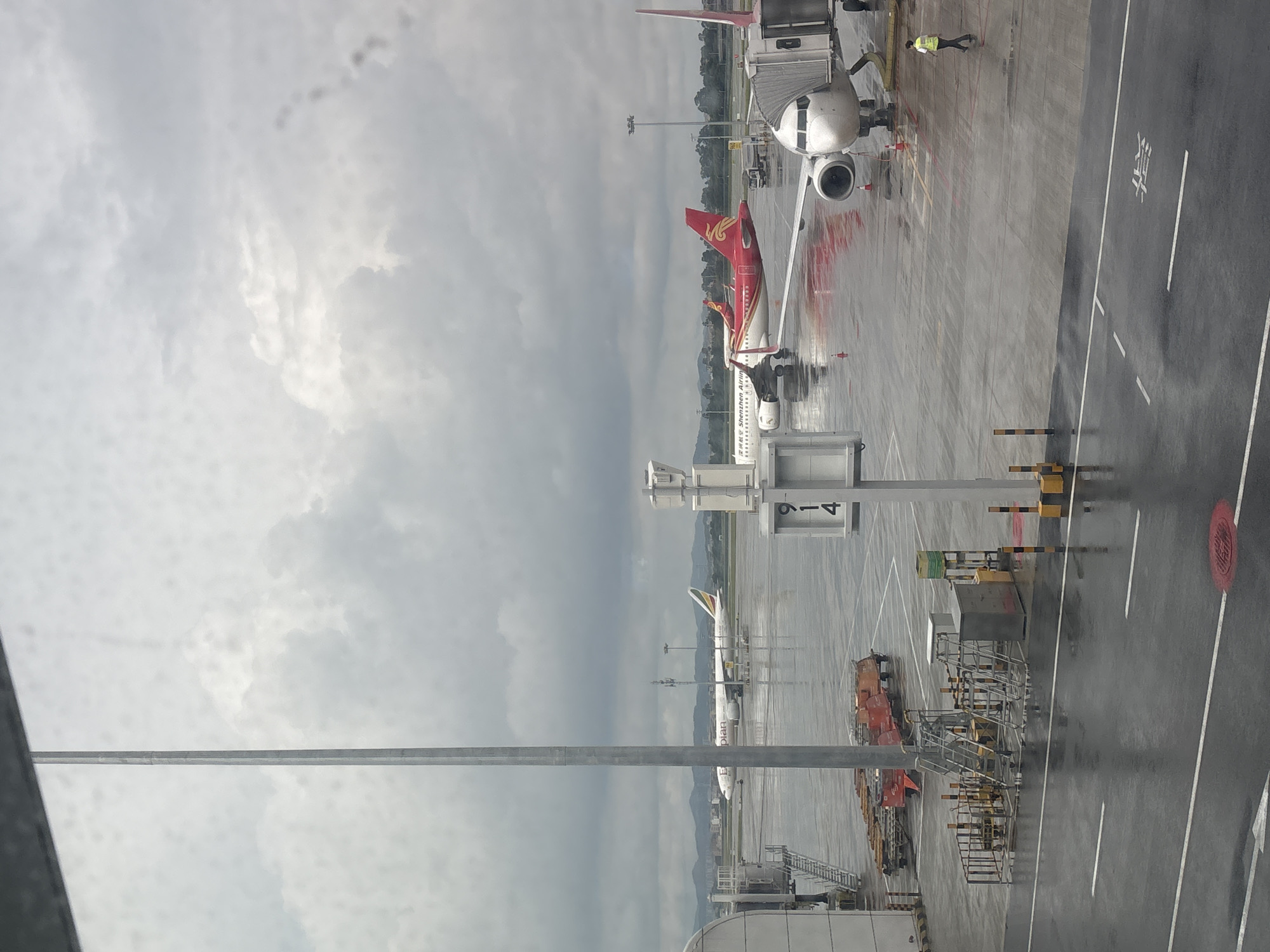 An airport tarmac is wet from rain, reflecting the overcast sky and the parked airplanes. In the foreground, a metal pole stands vertically on the left. A jet bridge extends from a terminal building on the far left. Several aircraft are visible on the tarmac. One is a white airplane with "ethiopian" written on its side, parked further back. In the middle ground, two red and white airplanes are parked. The airplane on the right, a Shenzhen Airlines aircraft, is closer to the foreground, connected to a jet bridge. A single person in a yellow vest and dark pants walks across the tarmac on the right side of the image. In the background, mountains are visible under a cloudy sky. Equipment such as carts and stairs are present on the tarmac.