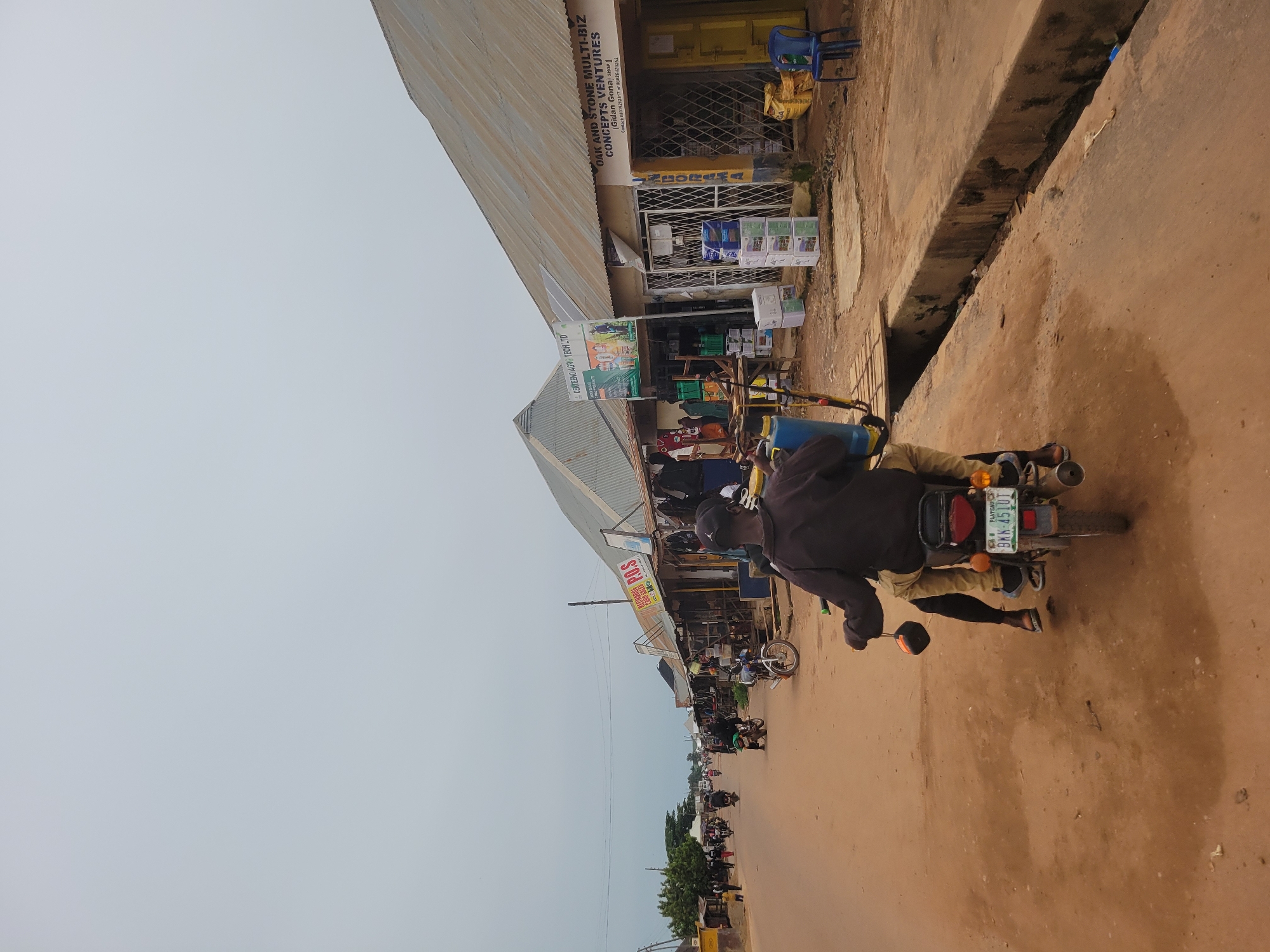 This is a sideways, eye-level, full-shot photograph taken in Bokkos, Nigeria. The scene depicts a roadside marketplace with several shops lining the street. The shops have corrugated metal roofs and are filled with various goods, including what appear to be agricultural supplies and electronic items.
A man is the primary subject, seen from behind riding a motorcycle down the dirt road. He is wearing a dark jacket and light-colored pants. The motorcycle has a license plate that reads "BKK-4510T" and "PLATEAU". Other motorcycles and a few people are visible further down the street, suggesting a busy day.
The sky is overcast and light, indicating it might be morning or late afternoon, and the weather appears to be neutral. The road is dusty and unpaved, typical of many areas in Nigeria. The overall atmosphere is one of everyday commerce and activity in a local market setting.
Visible text includes shop names such as "OAK AND STONE MULTI-BIZ CONCEPTS VENTURES," "CENTEENO AGRO TECH LTD," and signs indicating "RECHARGE CARD SALES" and "P.O.S." There are also contact phone numbers displayed on one of the shop signs. The image captures a slice of daily life in this Nigerian town.