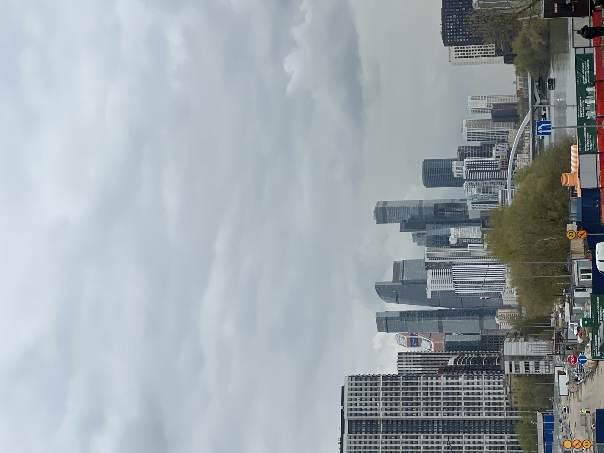 The image depicts a cityscape in Khimki, Russia, under an overcast sky. Multiple high-rise buildings form the skyline, including modern skyscrapers with varying architectural designs. Street-level views show signage including "STOP" and Russian text, indicating the presence of construction or road work. Utility poles, trees with sparse foliage, and hints of vehicular traffic are also visible. The environment suggests a mix of urban development and transportation infrastructure. A person is standing on the roadside.