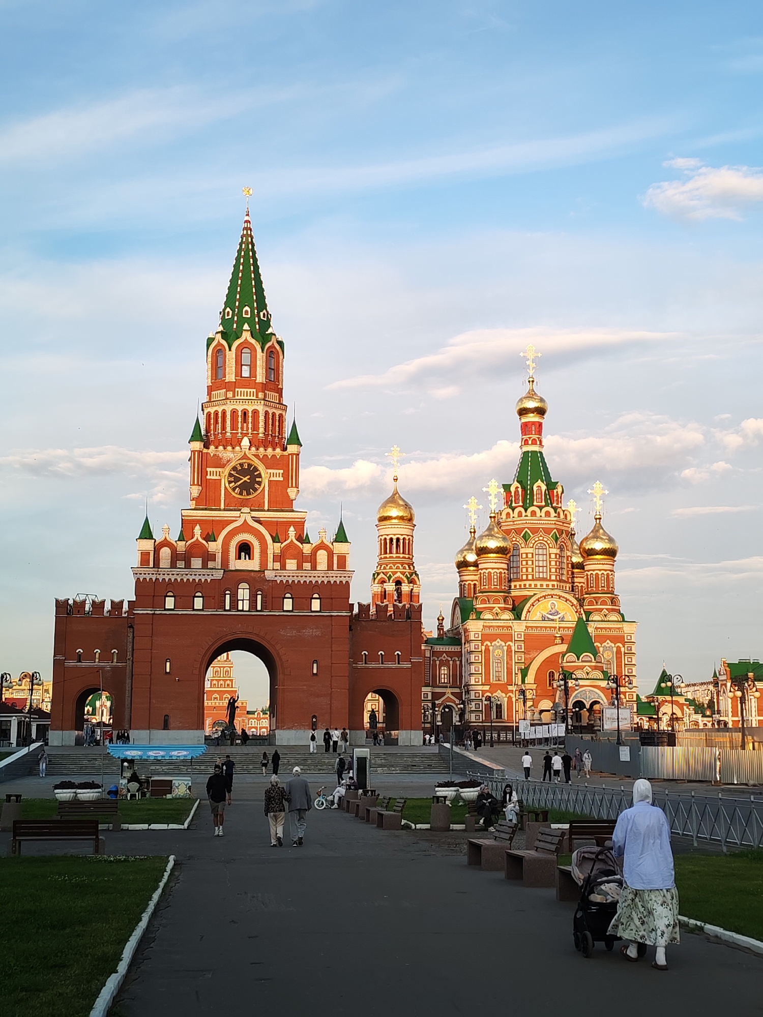 The image shows a wide shot of a public square in Yoshkar-Ola, Russia, featuring a prominent red brick tower with a green roof and a clock, adjacent to a brightly colored Orthodox cathedral with golden domes. Several people are walking through the square, including a woman pushing a stroller, an elderly couple, and other individuals in the background. The scene appears to be a public space with benches and grassy areas, suggesting it's a recreational or gathering spot.
The time of day is likely late afternoon or early evening, as indicated by the warm, soft light and the long shadows. The sky is partly cloudy with a soft blue hue, suggesting pleasant weather. The architecture of the buildings is ornate and distinctly Russian, with intricate patterns and vibrant colors. The tower resembles a Kremlin-style structure, and the cathedral is a classic example of Russian Orthodox church design.
The mood of the scene is peaceful and serene, with people enjoying a leisurely stroll in a beautiful, well-maintained public space. The presence of families and individuals of different ages suggests a community gathering or a popular tourist destination. There is no visible text in the image.