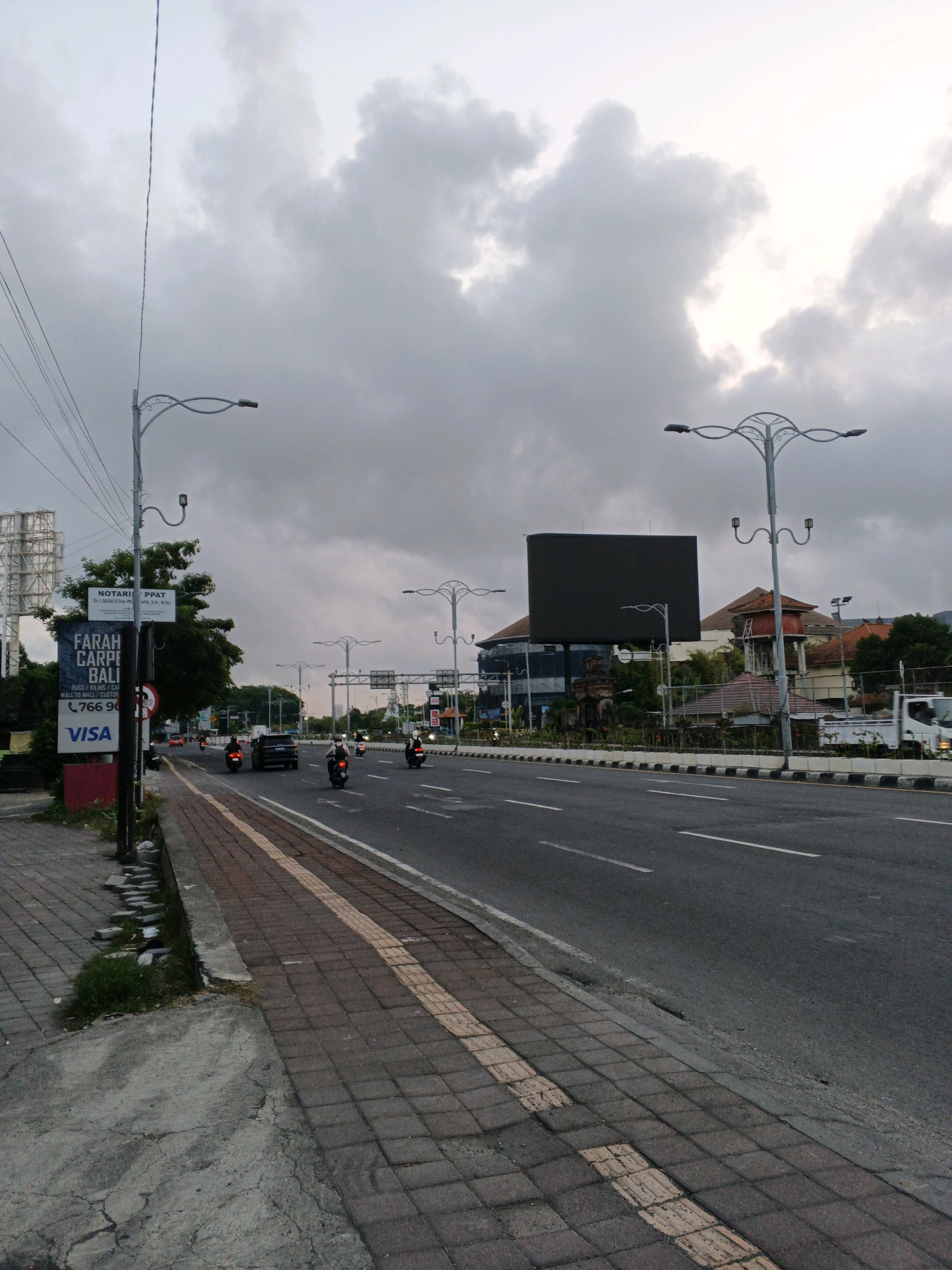 The image depicts a street scene in Kuta, Indonesia, under an overcast sky with heavy clouds. The view is from a sidewalk looking towards a multi-lane road with traffic. On the left side of the road, a sidewalk paved with interlocking bricks is visible, with some patches of grass and concrete. A signpost with several signs is also present, including one for "NOTARI PPAT" and another for "FARAH CARPET BALI" with a phone number and "VISA" logo.
The road itself has lane markings and several vehicles, including motorcycles and cars, in motion. Three motorcycles are clearly visible, with riders wearing helmets. A black car is also present. On the right side of the road, a row of decorative streetlights with multiple arms is installed, as well as power lines. In the background, buildings are visible, including one with a large, dark rectangular screen, possibly a digital billboard. Another building has a distinctive red water tower. The overall atmosphere is subdued due to the cloudy weather.