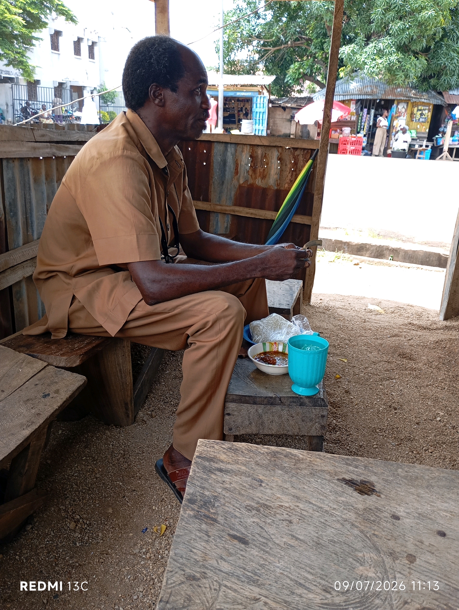 The image shows an outdoor scene in Shendam, Nigeria. A Black man, dressed in a brown uniform, sits on a wooden bench. He is looking to his right and holding a small object in his hands. In front of him, on another wooden surface, is a bowl of food, a plastic bag, and a turquoise cup filled with water. The background shows a market or roadside stall area with various structures and people in the distance. The ground is covered in small stones and sand. The lighting suggests it is daytime. The camera is positioned at a medium distance, capturing the man and his immediate surroundings.