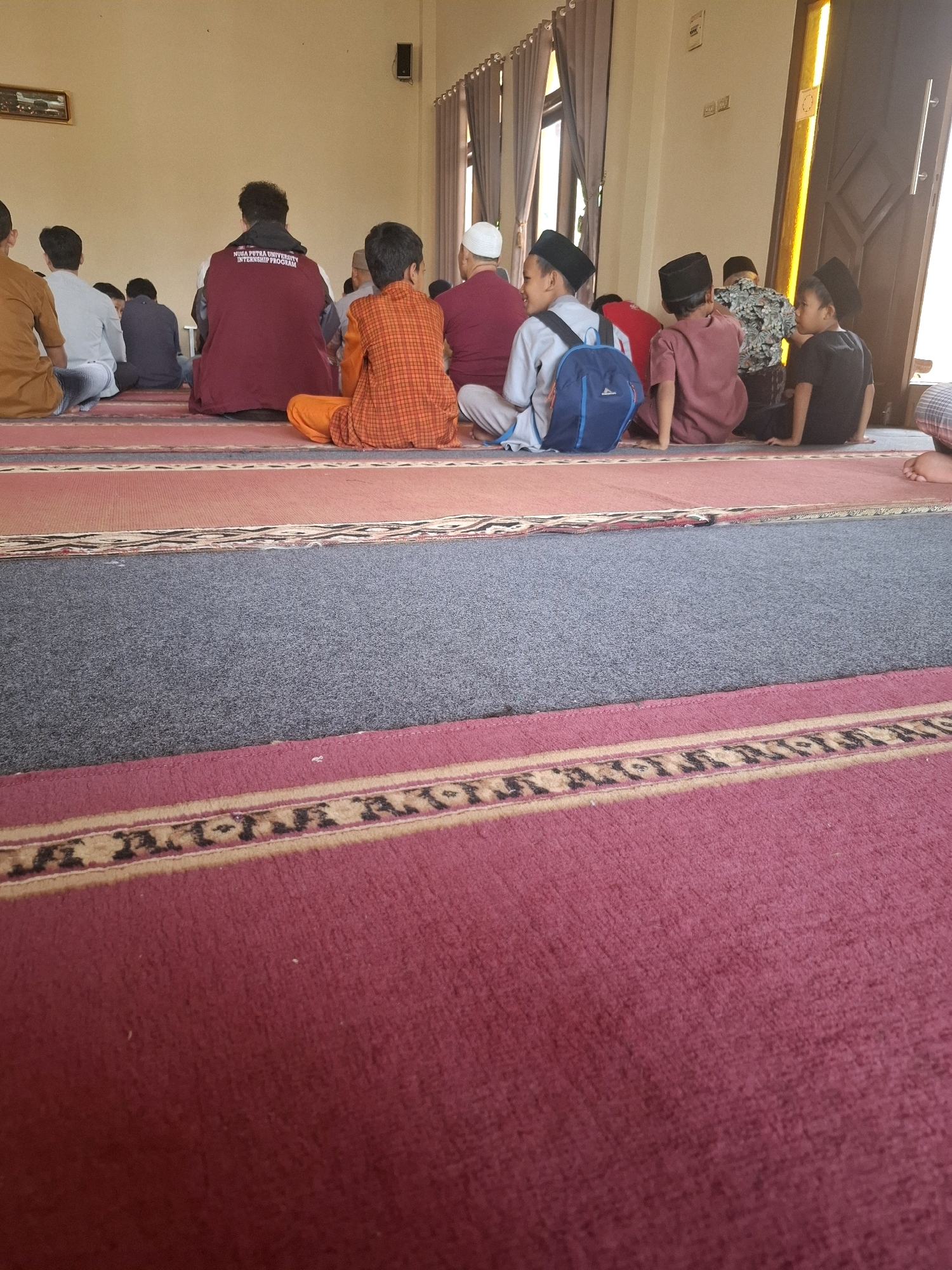 The image depicts a group of individuals, primarily young males, sitting on the floor inside what appears to be a mosque or prayer hall. They are facing away from the camera, towards a wall with curtains and a door. The floor is covered with carpets of varying colors and patterns, including red, gray, and a patterned stripe.
Several individuals are wearing traditional Islamic attire, such as prayer caps (peci) and long tunics. One person in the center foreground is wearing an orange checkered shirt and orange pants. Another person to their left is wearing a maroon sweatshirt with text that reads "NUSA PUTRA UNIVERSITY INTERNSHIP PROGRAM". Some individuals have backpacks. The lighting suggests it is daytime, with light coming from the right side of the frame, possibly from a window or doorway.
The overall atmosphere seems to be one of prayer or a religious gathering. The specific location is described as the city of Ciheulang Tonggoh, Indonesia. This detail is provided as context for the image.