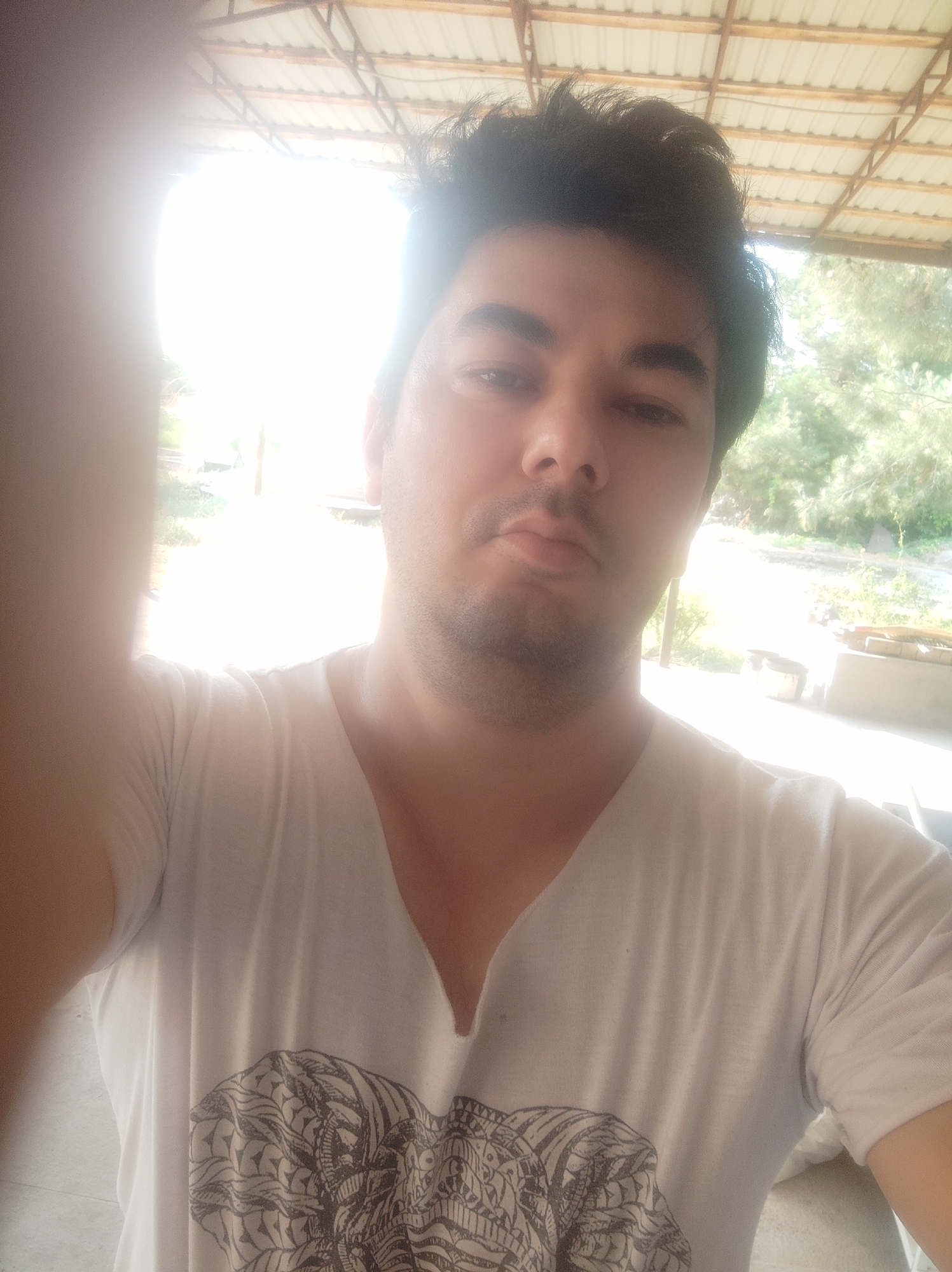 A selfie of a man wearing a white V-neck t-shirt with a black elephant graphic. He has dark hair and is outdoors. The background shows trees and a tin roof structure. The image is overexposed and hazy, making it difficult to discern specific details. The man is positioned in the foreground, with his left arm blurred and covering part of the frame. The location is identified as Mingchinar (Blagoveshchensky), Uzbekistan.
