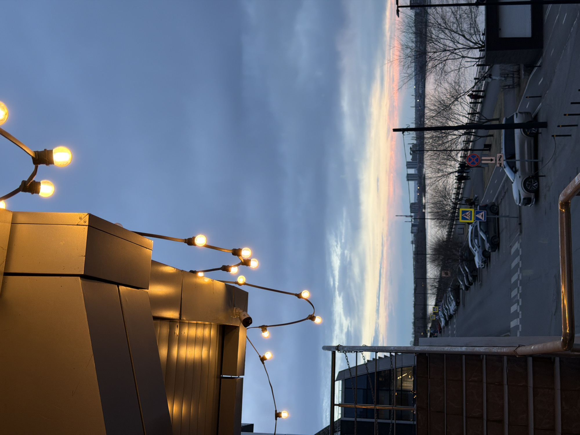 The image depicts a twilight scene in Krasnoyarsk, Russia. A view of the Yenisey River is visible in the background, with a strip of orange sunset light present just above the treeline. Along the riverbank, a road is lined with parked cars, streetlights, and traffic signs. A pedestrian crosswalk is present. In the foreground, architectural elements of a building are adorned with string lights. A metal staircase and railing are also visible.