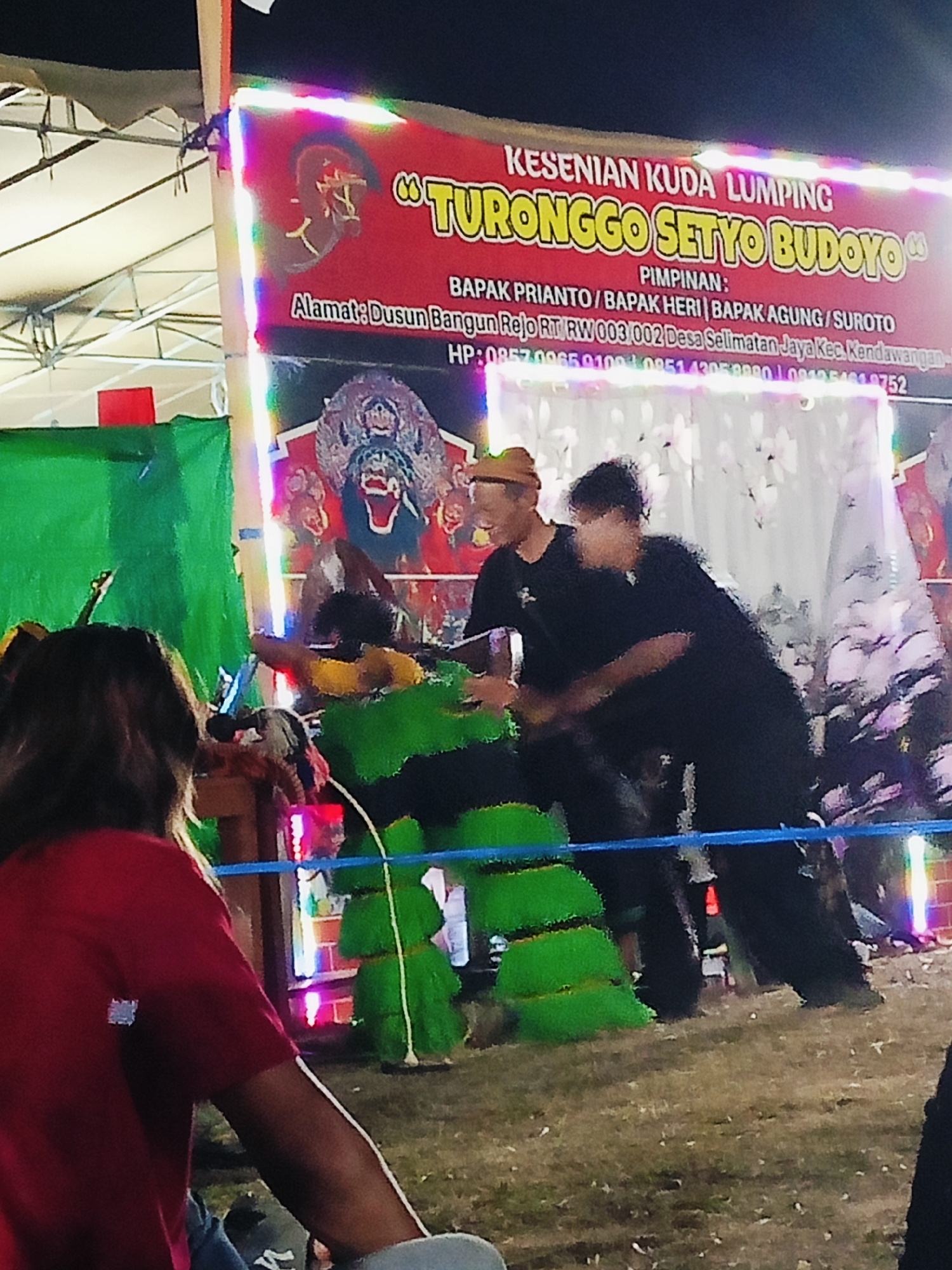 The image shows an outdoor nighttime performance of Kuda Lumping, a traditional Indonesian dance, at the location of Dusun Rejo, RT RW 003 002, Desa Selimantan Jaya, Kecamatan Kendal, Kabupaten Kendal, Indonesia. A red banner with illuminated text stating "KESENIAN KUDA LUMPING TURONGGO SETYO BUDOYO" is prominently displayed behind the performers. The banner also lists the leaders and contact information for the group, including phone numbers. In the foreground, a person in a red shirt is seated, facing away from the camera, towards the performance. The performance itself involves two individuals in black shirts interacting with a green, horse-like effigy, possibly symbolizing the "kuda lumping." The ground appears to be a dirt or grass field. The overall scene is illuminated by the banner's lights and possibly other ambient lighting from the event.