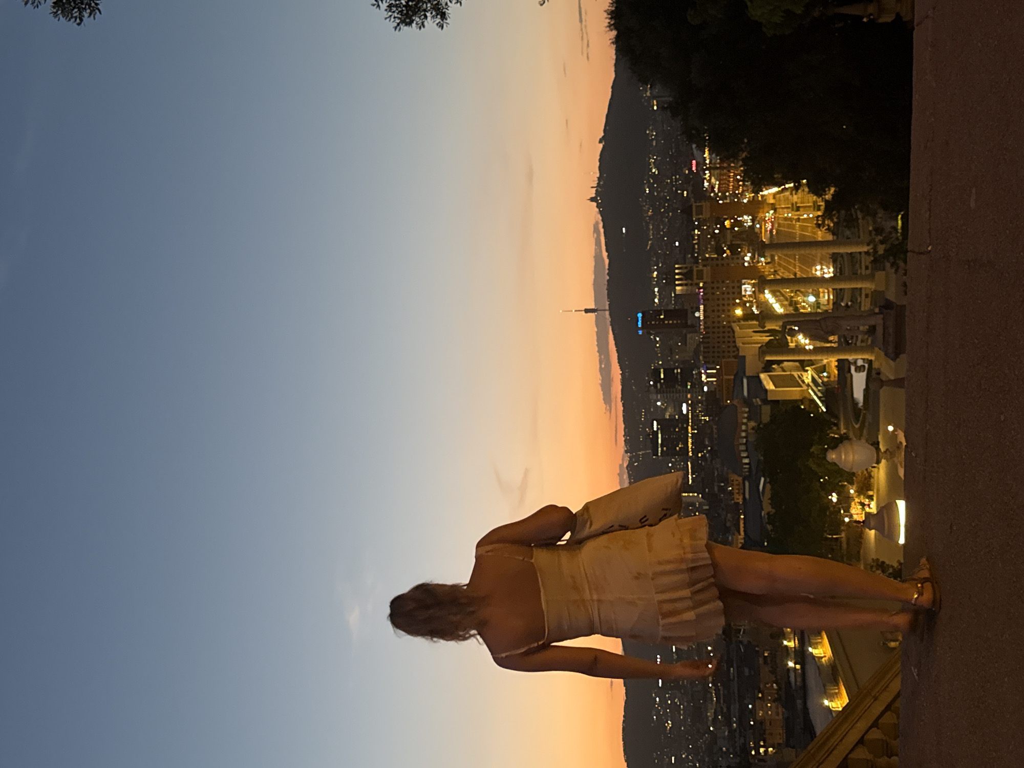 A blonde woman stands on a balcony overlooking the city of Barcelona at dusk. She is wearing a light-colored, ruffled dress and sandals, and carries a tote bag over her shoulder. The city lights are beginning to twinkle below, and the sky is a gradient of blue and orange. In the distance, a hill with a few buildings is visible. The woman is facing away from the camera, gazing at the panoramic view. The scene captures a moment of quiet contemplation as the day turns into night. The location is likely from a high vantage point, such as Montjuïc hill, which offers expansive views of the city.