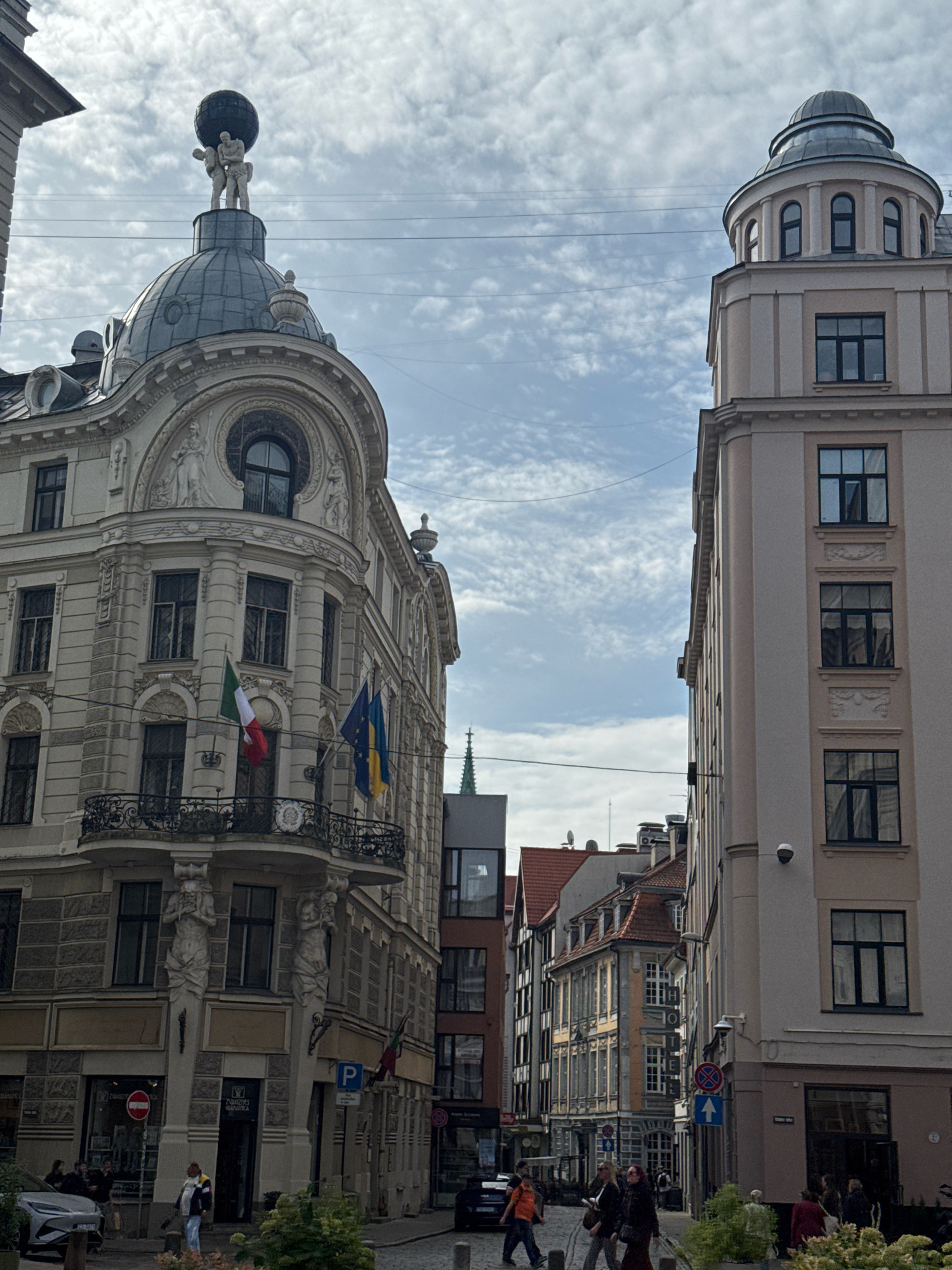 The image depicts a street scene in Riga, Latvia, featuring ornate historical architecture. In the foreground left, a cream-colored building with intricate carvings and a dome-topped structure stands prominently. A sculpture of two figures carrying a globe adorns the apex of this dome. Two flags, one Italian and one possibly Ukrainian, are displayed on a balcony of this building. To the right, a taller, lighter-colored building with a rounded turret and arched windows rises. The street itself is paved with cobblestones, and several individuals are visible walking, with some gathered near shop entrances. The sky is partly cloudy. The overall impression is of a well-preserved European city with a blend of architectural styles.