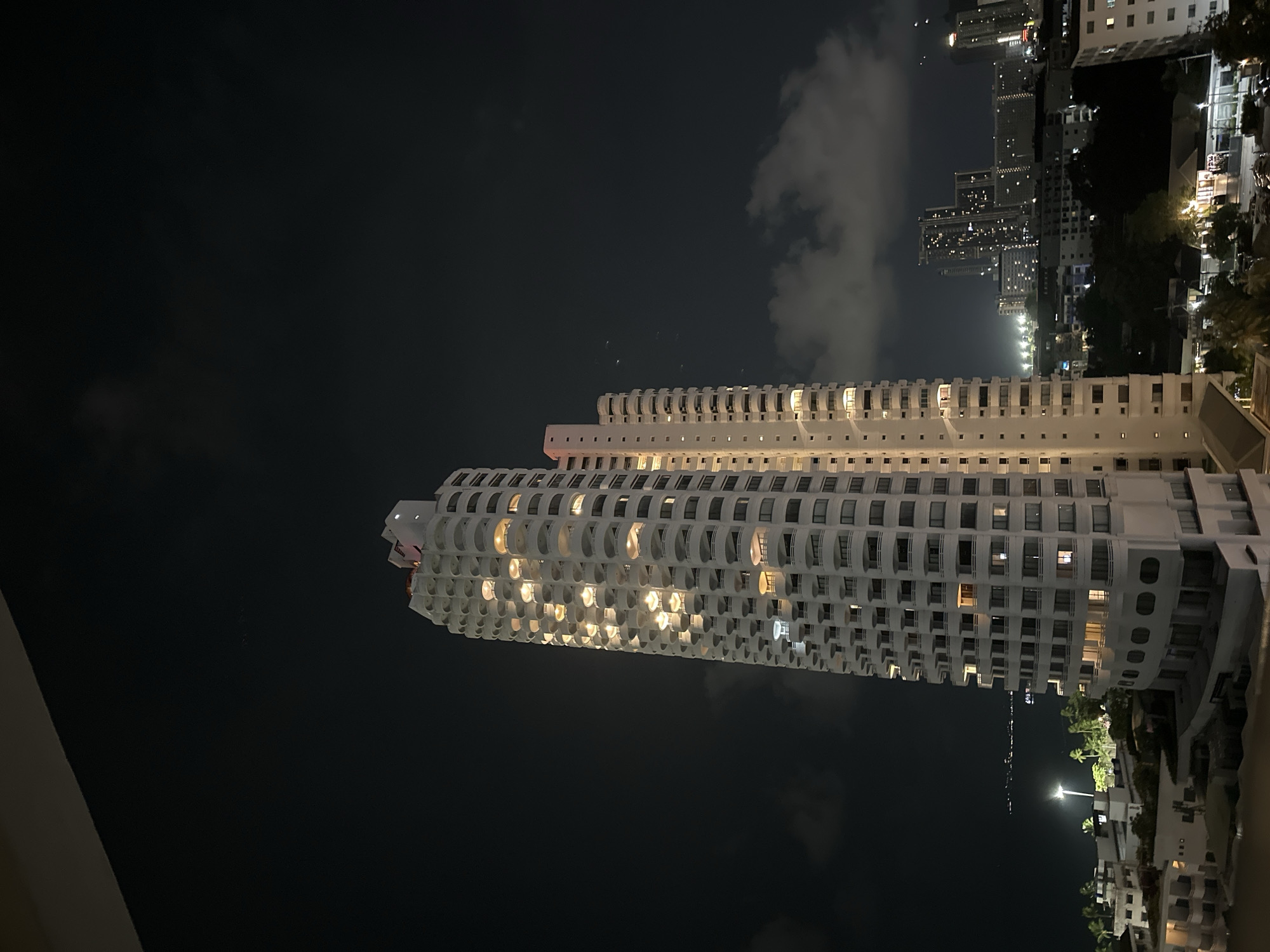 At night, the city of Pattaya City, Thailand, is viewed with many towering buildings. The foreground features the Dusit Thani hotel. The building has lights on in the windows, creating a warm glow. The buildings in the background are also illuminated, and the sky is dark with some clouds.