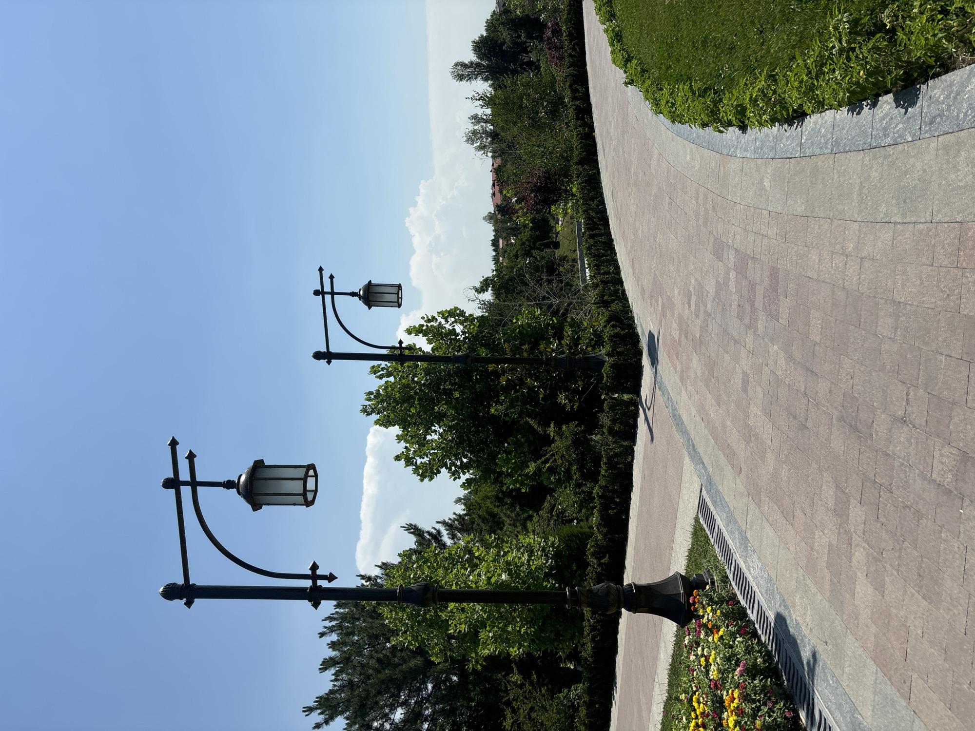 The image depicts a park with two ornate black lampposts lining a paved walkway. The walkway is made of light brown rectangular tiles and curves to the right, bordered by neatly trimmed green bushes on the right and a flowerbed with colorful flowers on the left. In the background, a dense green forest with various trees and some buildings are visible under a clear blue sky with scattered white clouds. This scene is located in Tashkent, Uzbekistan, specifically at the Alley of Wisdom.