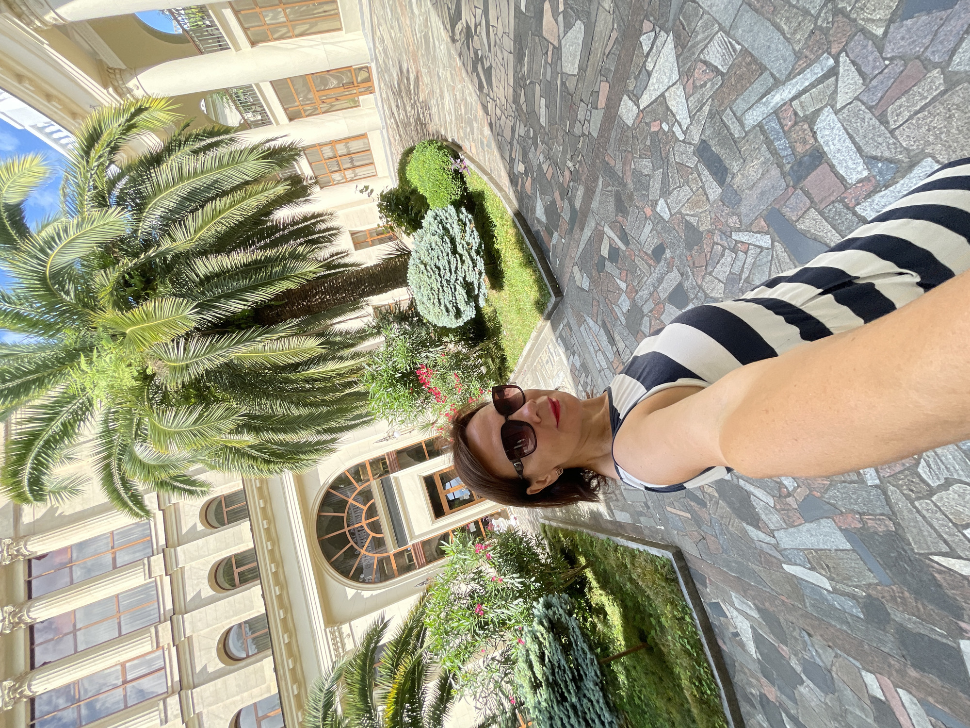A woman is taking a selfie in a courtyard in Gagra, Abkhazia. She is wearing a black and white striped dress and sunglasses. The courtyard is paved with colorful stones and surrounded by greenery, including palm trees and bushes. A large, ornate building with columns and arched windows is visible in the background. The weather appears sunny.