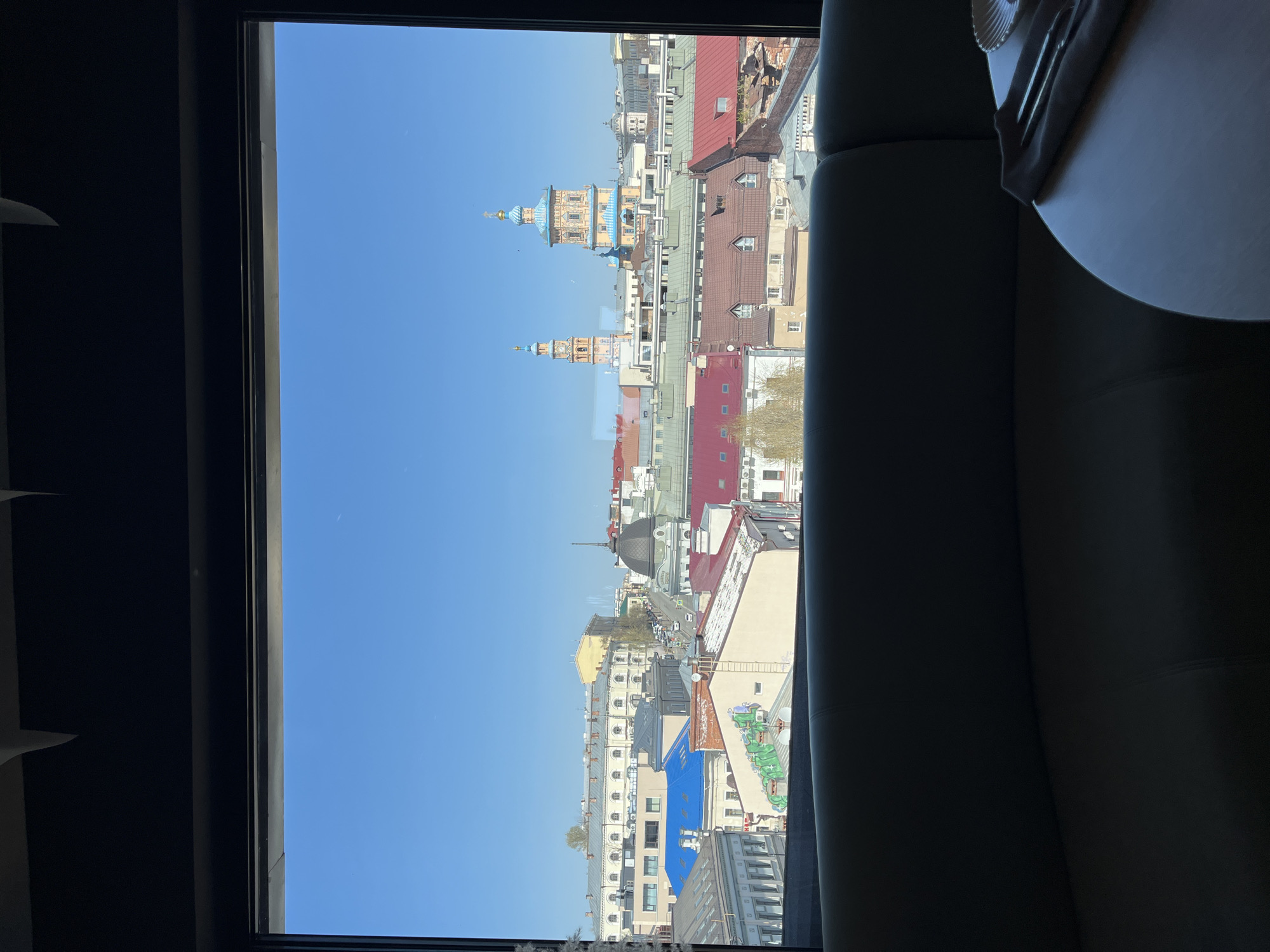 The image depicts a view of Kazan, Russia, from an interior window. The cityscape includes buildings of various heights and colors, including red, blue, and tan. Two notable buildings with domed towers are visible against a clear blue sky. To the left of the window is a dark wall. To the right of the window, part of a piece of furniture is visible.