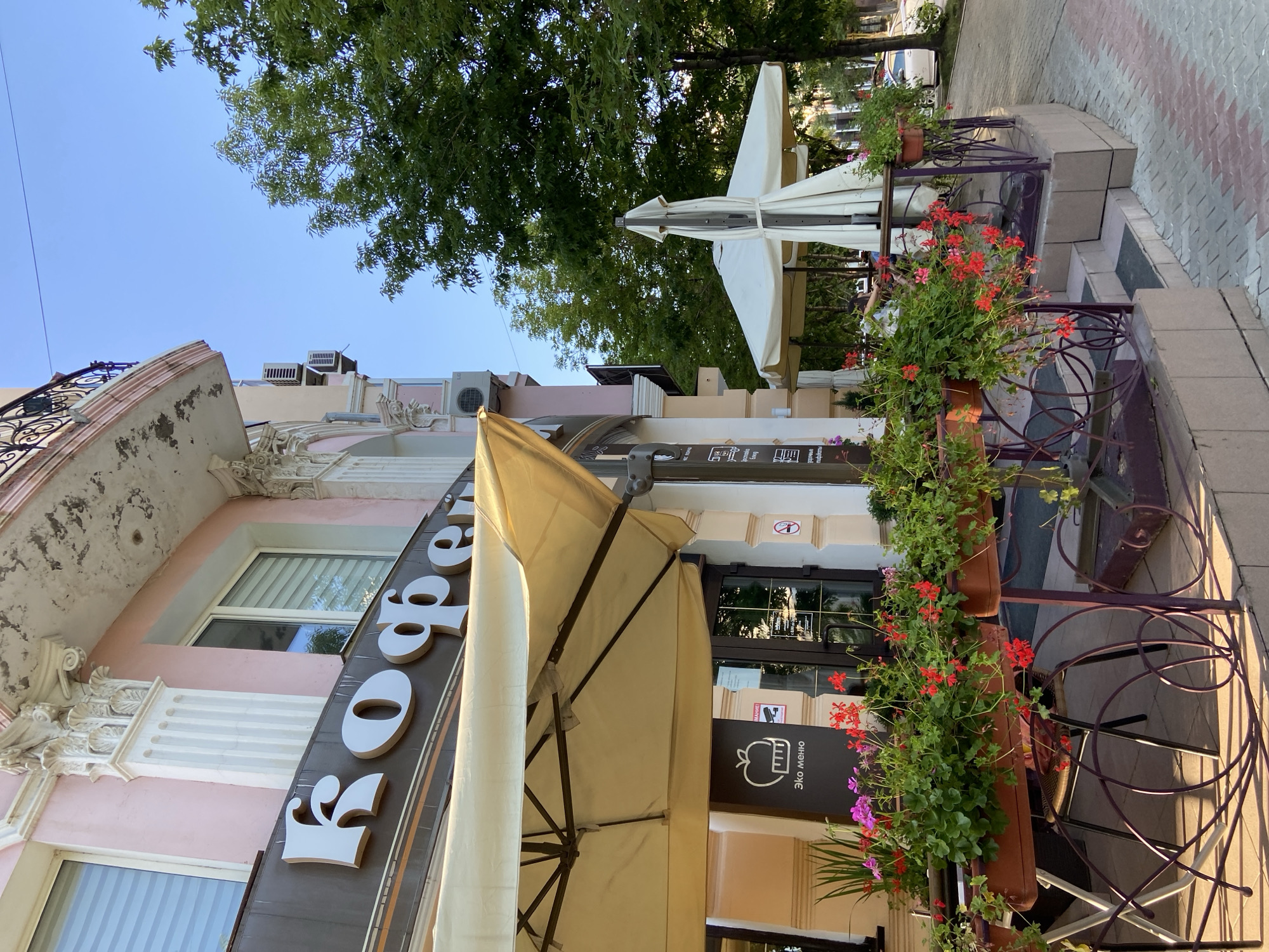 A low-angle, close-up exterior shot shows the facade of a pink building housing a cafe in Blagoveshchensk, Russia. The sign above the cafe entrance reads "Кофе" (Coffee) in large white 3D letters. An open beige umbrella shades an outdoor seating area where potted flowers in shades of pink and red bloom. Another umbrella, closed and tied, stands nearby. A dark sign with a white apple logo and text reads "Эко меню" (Eco menu). To the right of the building, green trees line a sidewalk, and a white car is parked on the street. The building's architectural details, including ornate white trim and decorative columns, are visible, as is some peeling paint on the upper facade and balcony. The sky above is a clear blue.