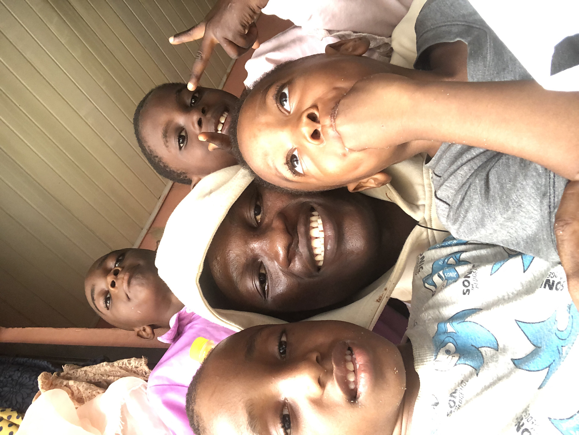 The image is a selfie taken outdoors in Ado, Nigeria. In the foreground, a Black man wearing a hooded sweatshirt is smiling broadly, with five Black children in various states of looking at the camera or away from it. The children are positioned around the man, with some partially obscured. The youngest child, in the front right, has their fist in their mouth. The environment appears to be covered, with a slatted ceiling visible in the background. There are also hints of patterned fabric in the upper left corner, possibly curtains or clothing. The lighting suggests it is daytime.