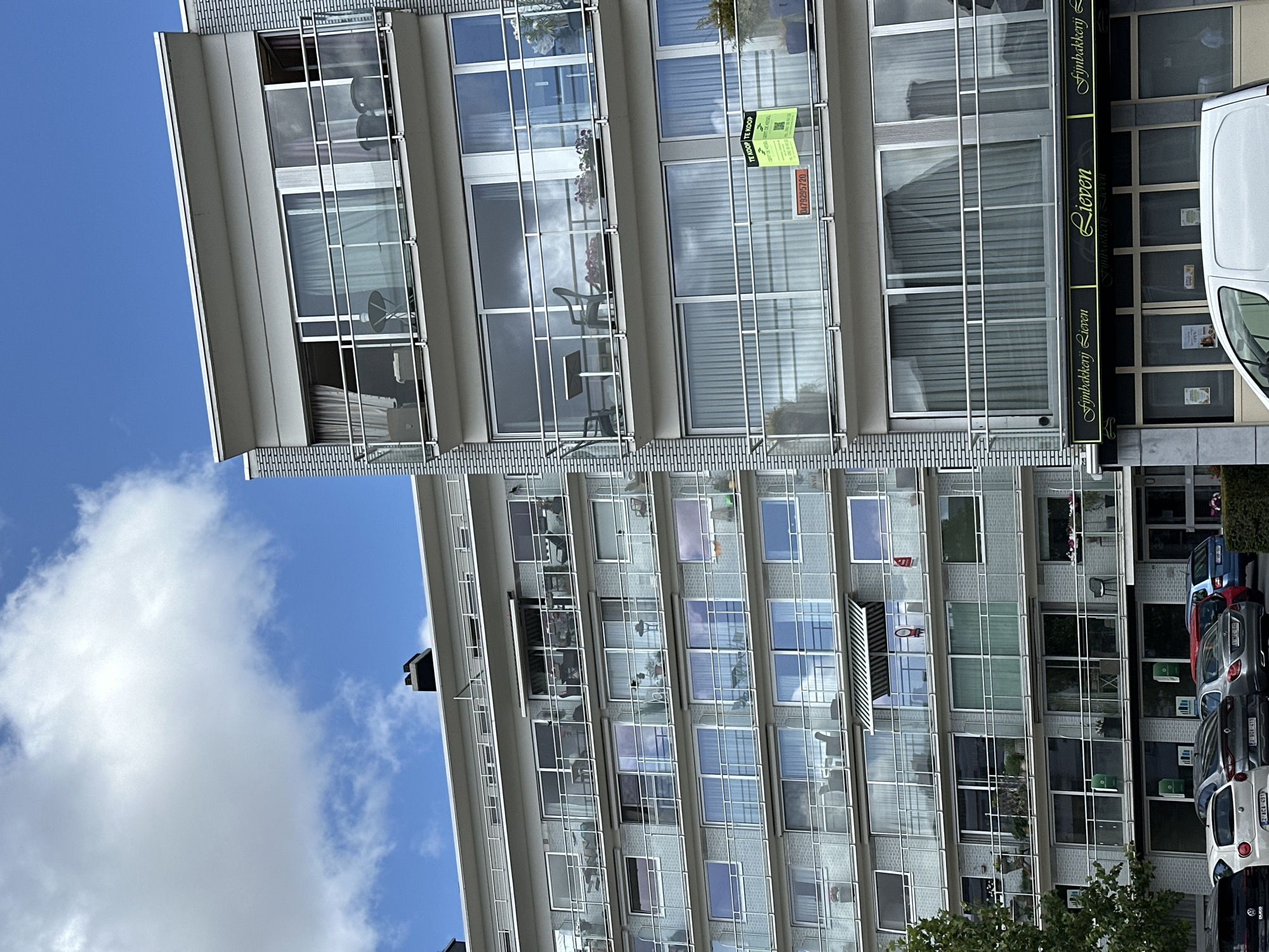 The image shows a multi-story modern apartment building in Bruges, Belgium. The building has numerous balconies with glass railings and sliding doors leading to them. Some balconies are furnished with chairs and tables. On one of the balconies, a "TE KOOP" (For Sale) sign is visible. The ground floor of the building houses a bakery with a sign that reads "Fijn Bakkerij Lieven" and "Lieven." Several cars are parked in front of the building. The sky is blue with scattered clouds, and a tree is partially visible on the left side of the frame. The architecture is contemporary with a light-colored brick facade interspersed with metal elements.