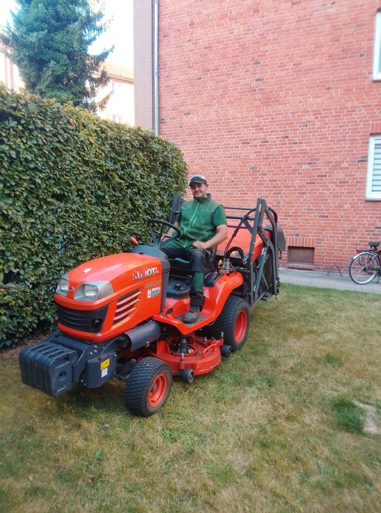 A man wearing a green vest over a dark shirt and pants, and a dark baseball cap, is sitting on an orange Kubota G231 ride-on mower. The mower is positioned on a grassy lawn in front of a tall, dense green hedge. Behind the hedge is a brick building with several windows. To the right of the building, a bicycle with a red frame and black wheels is visible. The man is looking towards the camera with a slight smile. The mower has a black front bumper, a steering wheel, and a seat. The grass appears somewhat dry in patches. The scene is outdoors, likely in a residential or park area.