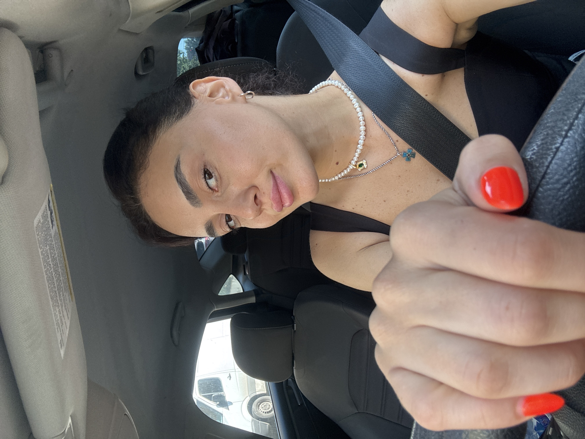 A young woman with dark hair and brown eyes, wearing a black tank top and a pearl necklace, sits in the driver's seat of a car. She has bright red nail polish on her fingers and is holding the steering wheel. The interior of the car is visible, with black seats and a seatbelt across her chest. Outside the car window, a white van is parked. The scene appears to be a street in Tbilisi, Georgia.