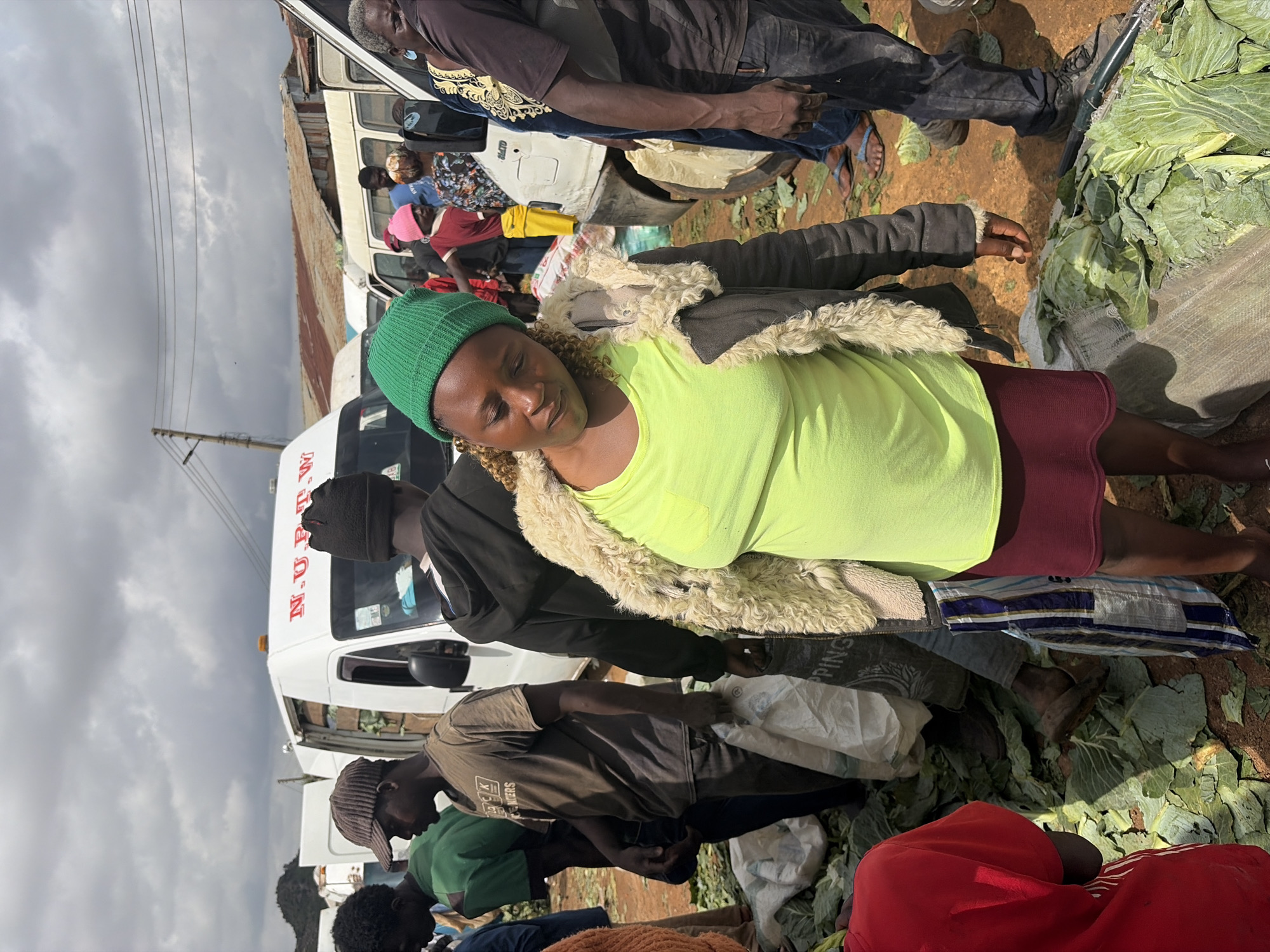 An outdoor marketplace scene in Gura Top, Nigeria, shows a group of people surrounded by large piles of leafy green vegetables, likely cabbage. In the foreground, a woman wearing a bright green beanie and a neon yellow top with a fluffy-collared jacket looks down, her arms slightly bent. She is standing next to a dark red skirt. To her left, a man in a brown shirt and a dark baseball cap is carrying a white plastic bag. Behind him, a man in a green shirt is also present. Several white vans are parked in the background, one with "N.U.R.T.W" visible on its side. The ground is covered in dirt and scattered vegetable leaves. The sky is cloudy.